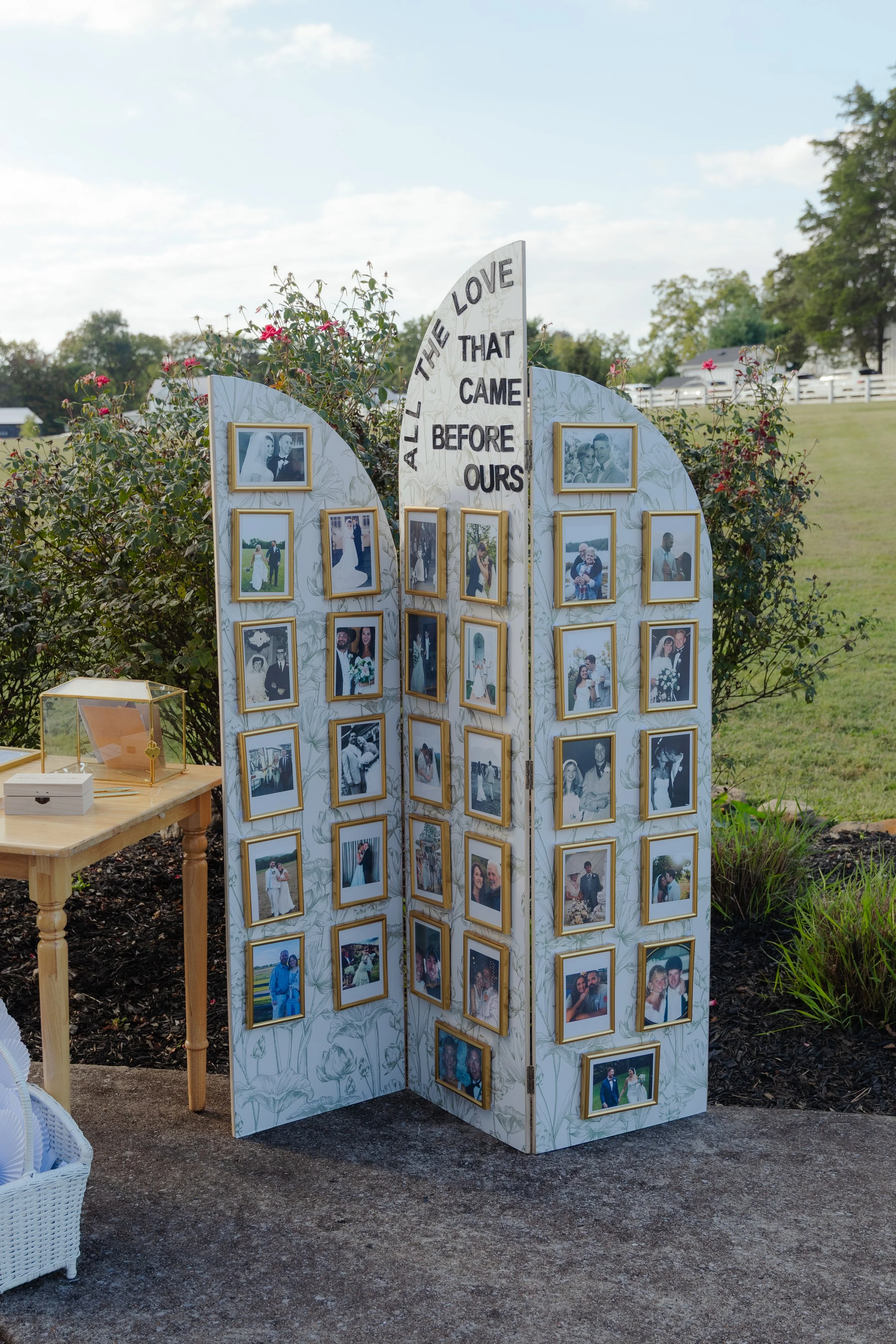 A photo display of framed wedding photos on a folding screen with the phrase 'ALL THE LOVE THAT CAME BEFORE OURS' at a wedding reception outdoor in daytime, with greenery and blue sky in the background.
