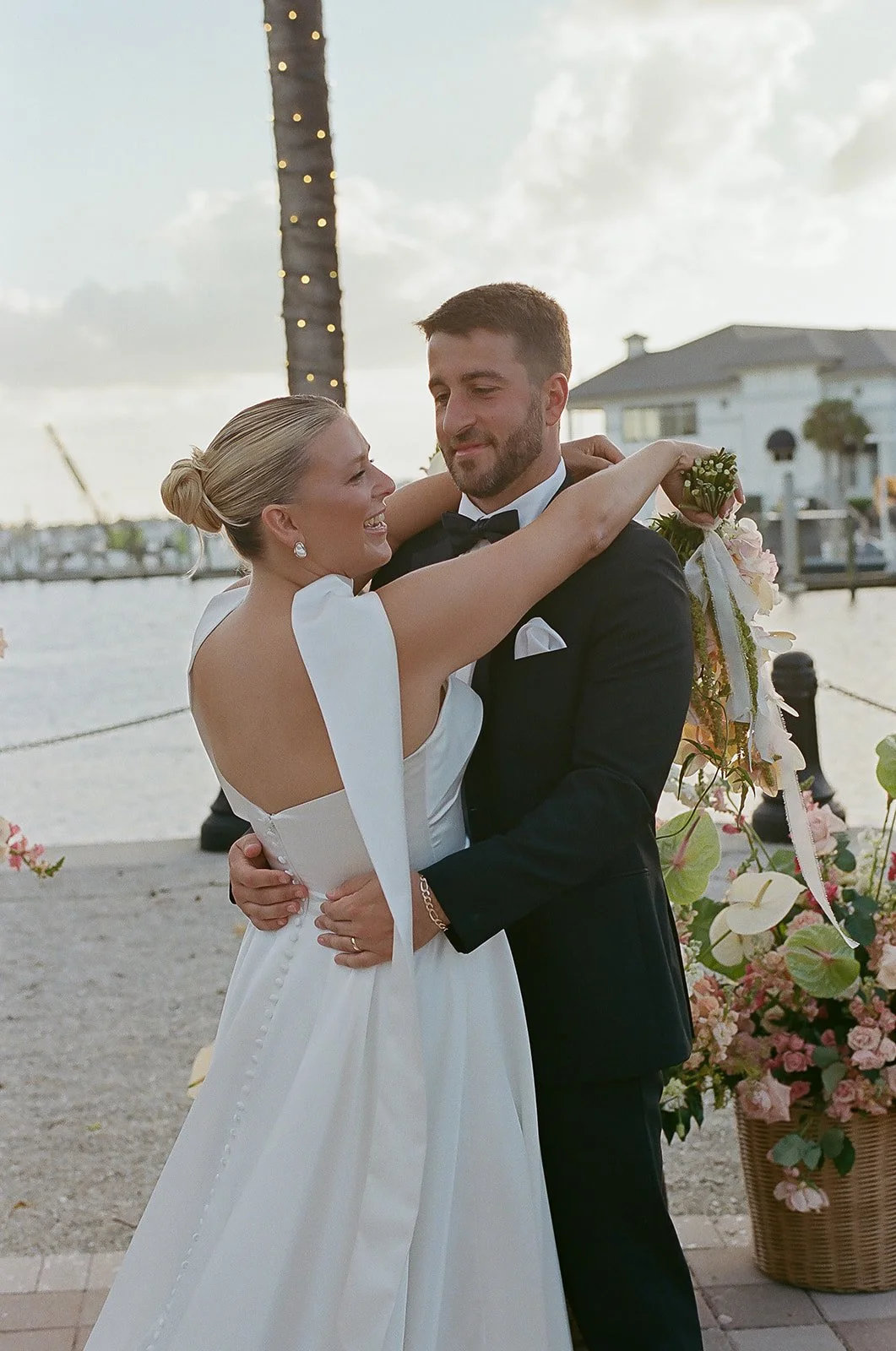 A bride and groom embrace during their wedding reception outdoors near a body of water, with decorative flowers and a lit palm tree in the background.
