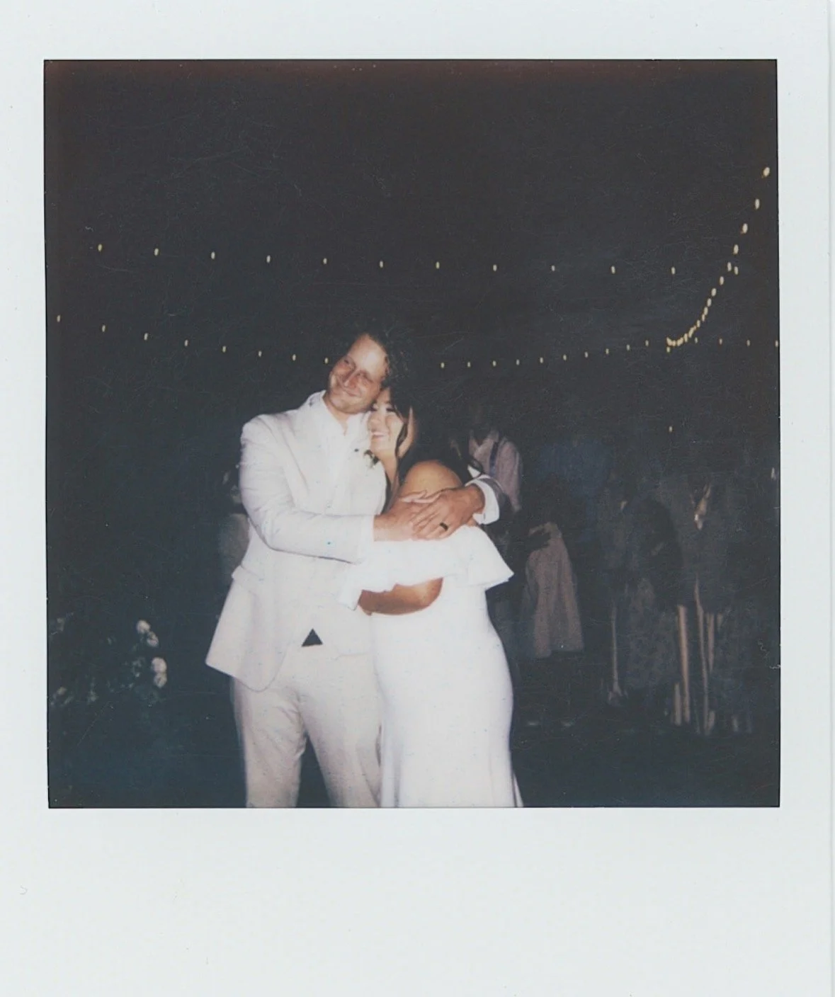A bride and groom dancing at their wedding reception, smiling and hugging each other under string lights at night.