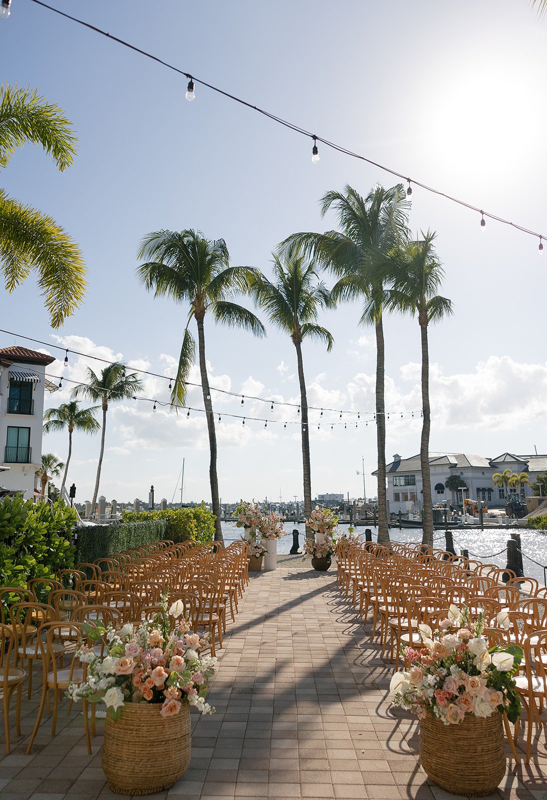 Outdoor wedding setup at a waterfront with rows of chairs, flowers in woven baskets, palm trees, string lights, and a marina in the background.