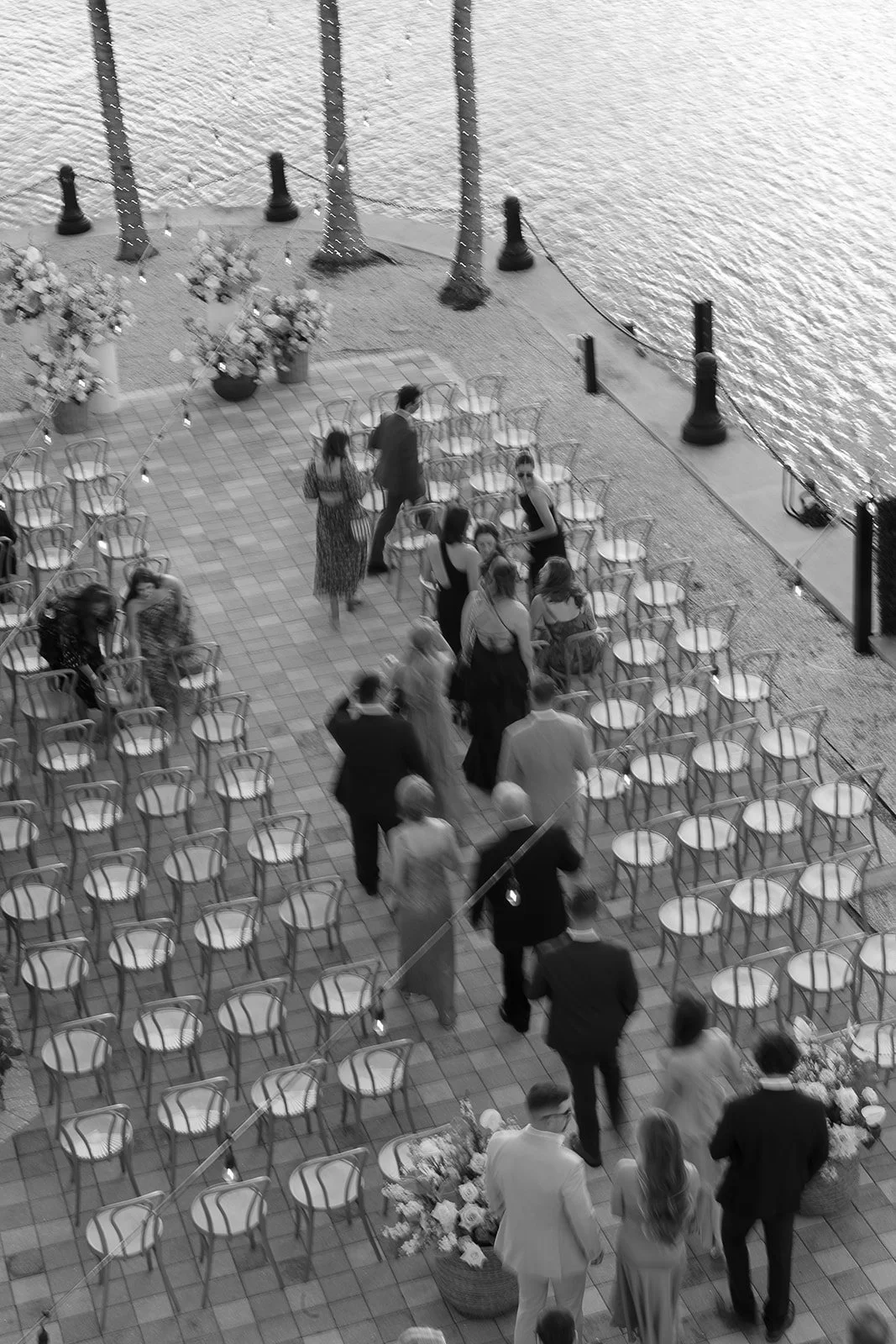 A group of people dressed in formal attire gathered on an outdoor terrace overlooking a body of water, possibly for a wedding or special event. The area has tables and chairs, with some chairs arranged in rows. There are large flower arrangements on 