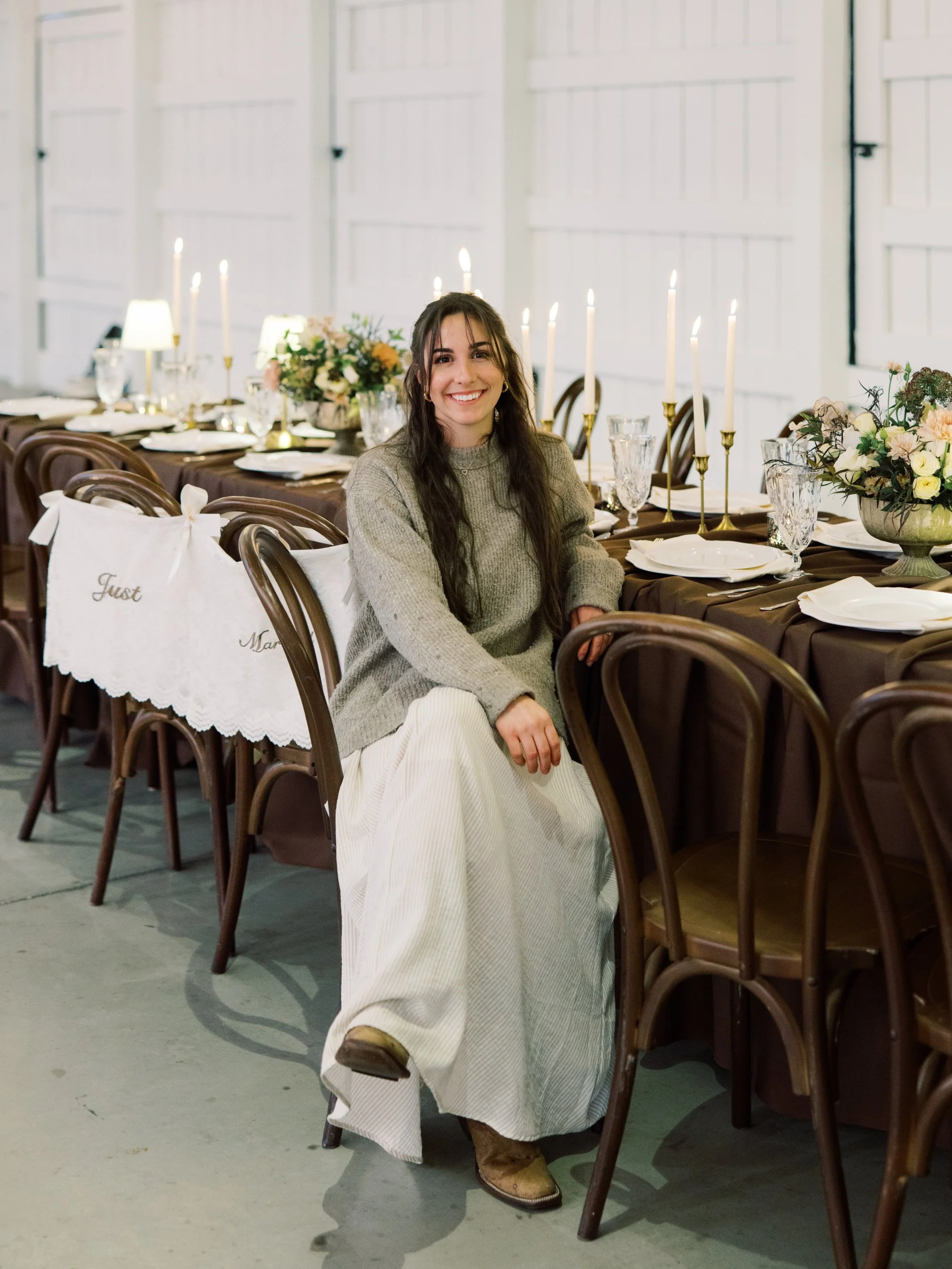 A woman sitting at a decorated dining table with candles and floral arrangements, smiling at the camera.