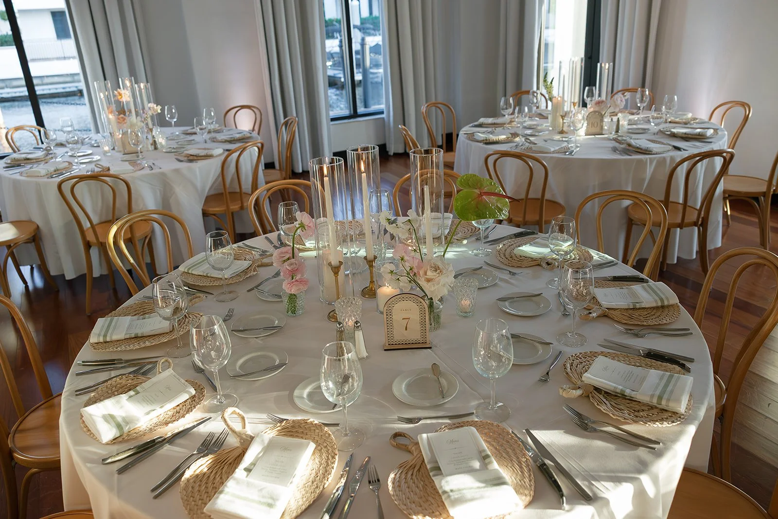 Round banquet tables set with white tablecloths, glassware, cutlery, napkins, woven placemats, and floral centerpieces in a well-lit room with large windows and wooden flooring.