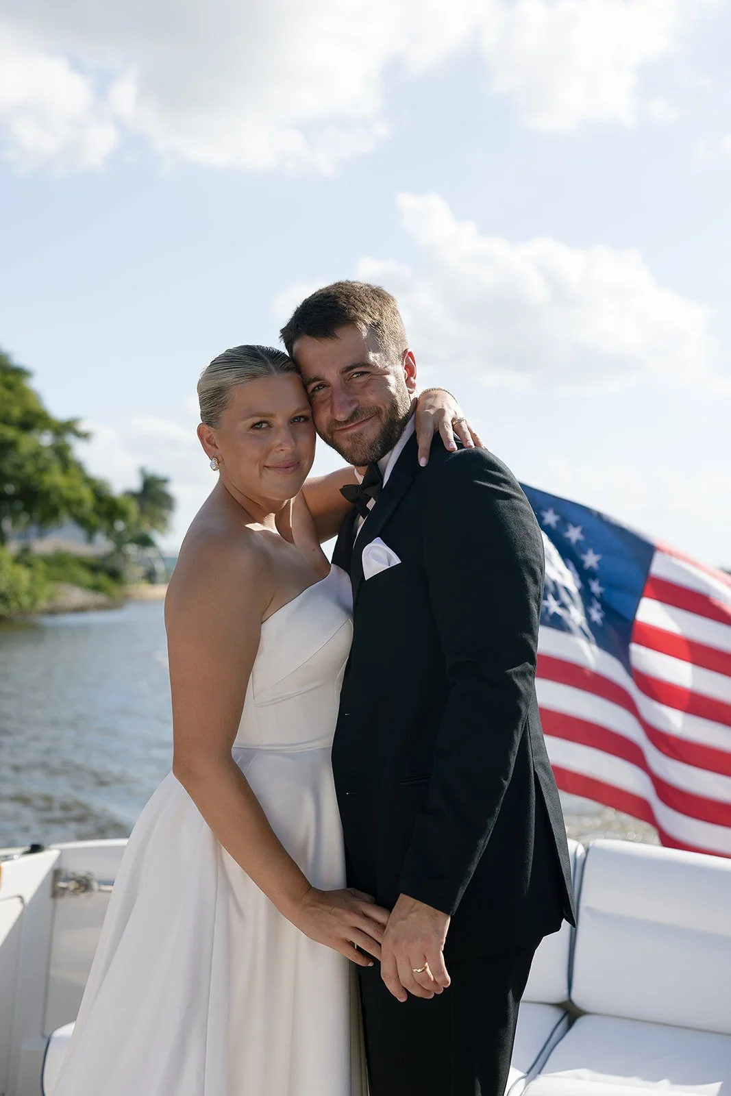 A newlywed couple in wedding attire embraces on a boat with an American flag in the background, near a body of water with trees and a cloudy sky.
