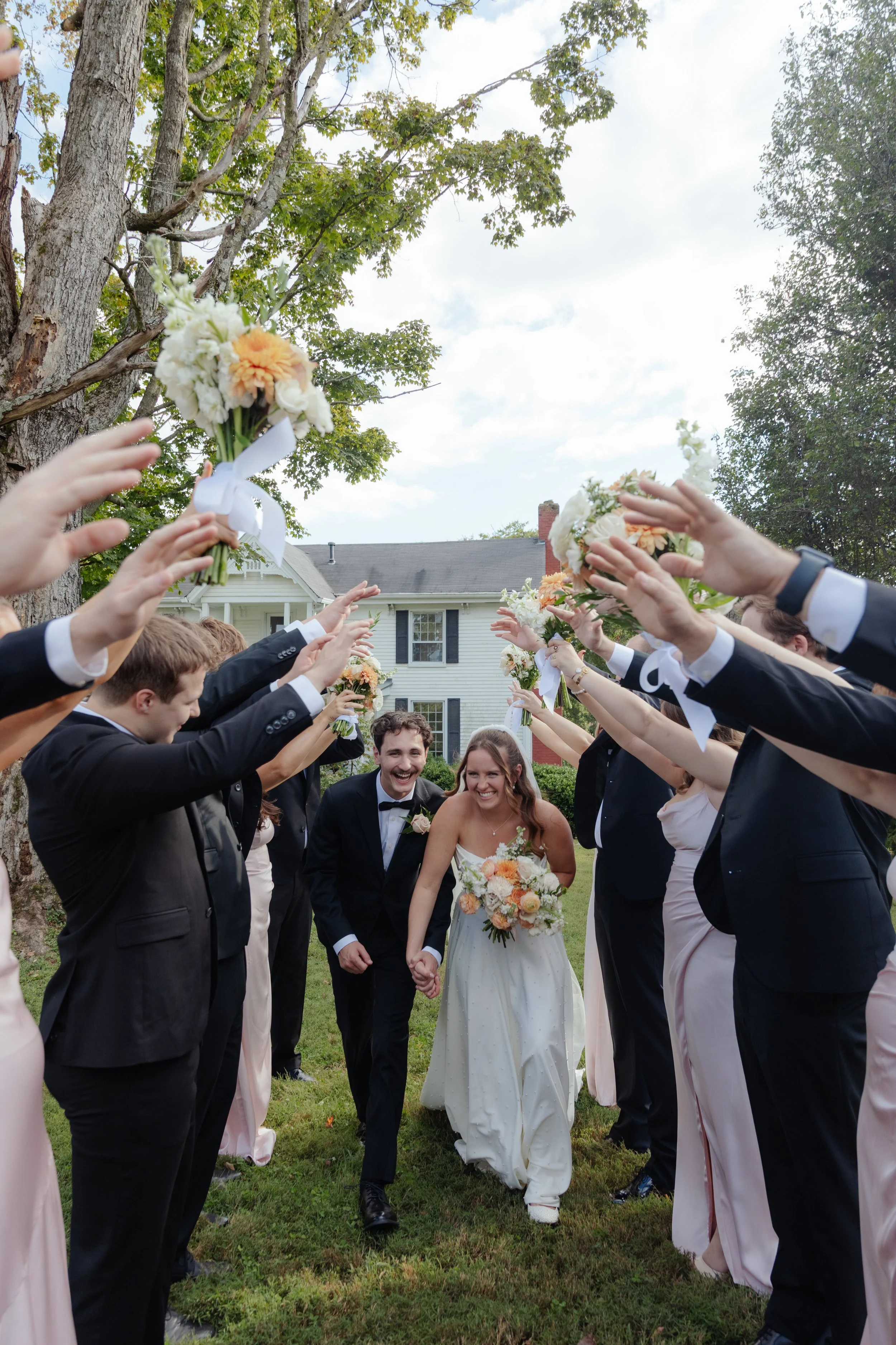 A wedding celebration scene with a bride and groom walking hand-in-hand through an archway created by guests holding bouquets of flowers, outdoors on a grassy lawn with trees and a house in the background.