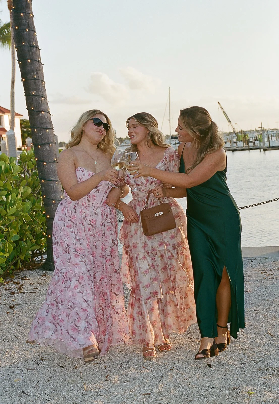 Three women in elegant dresses raising wine glasses in a toast by the waterfront, with boats and a dock in the background.