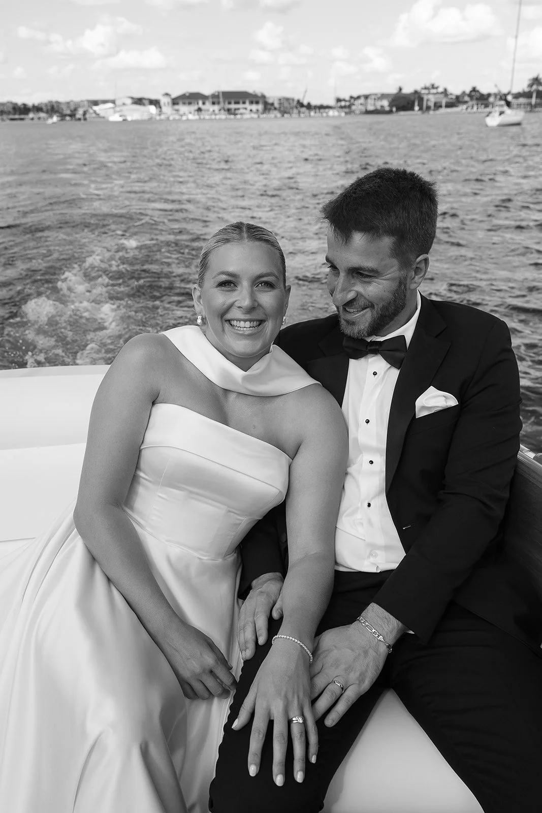 A smiling bride and groom sitting on a boat by the water, dressed in wedding attire, with a background of water and buildings.