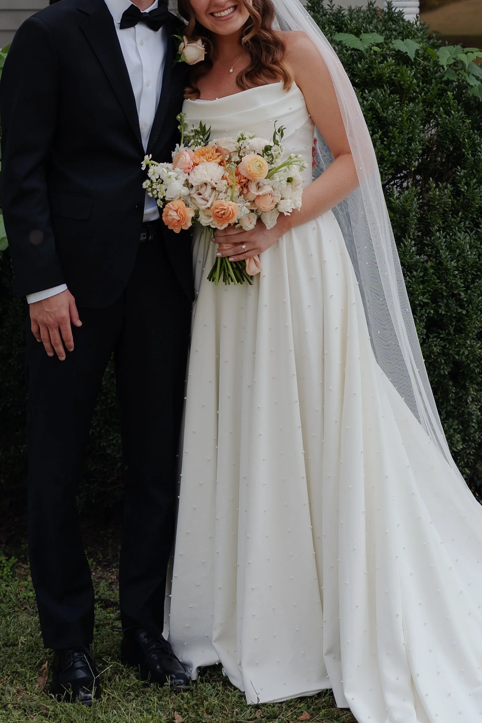 A bride and groom standing together outdoors on their wedding day, with the bride holding a bouquet of flowers and wearing a white wedding dress and veil, and the groom in a black tuxedo with a bow tie.