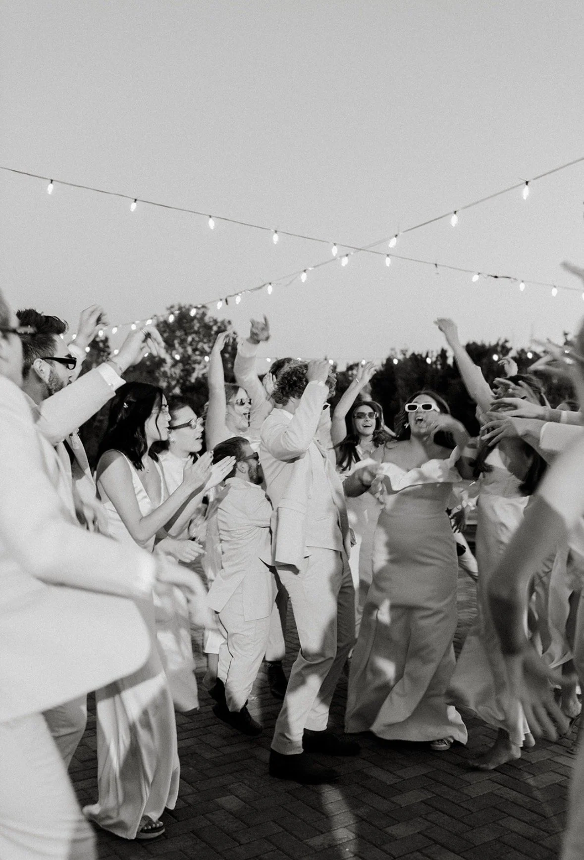 Group of people dancing outdoors at night under string lights, dressed in formal attire, with some wearing sunglasses.