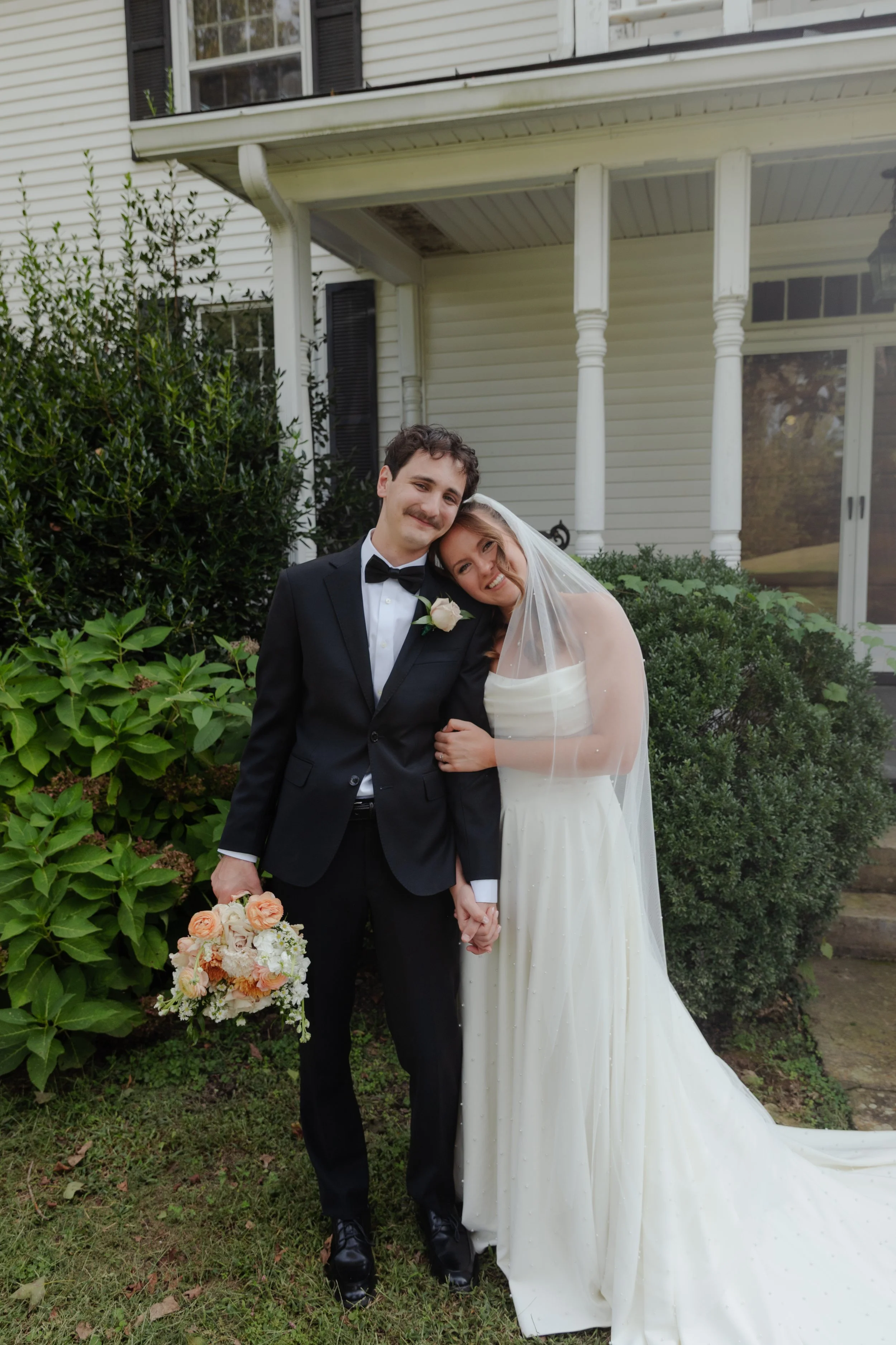 A newlywed couple stands outdoors in front of a house, smiling and holding hands. The groom is dressed in a black tuxedo with a bow tie and is holding a bouquet of flowers. The bride is wearing a strapless wedding gown and a veil, leaning her head on