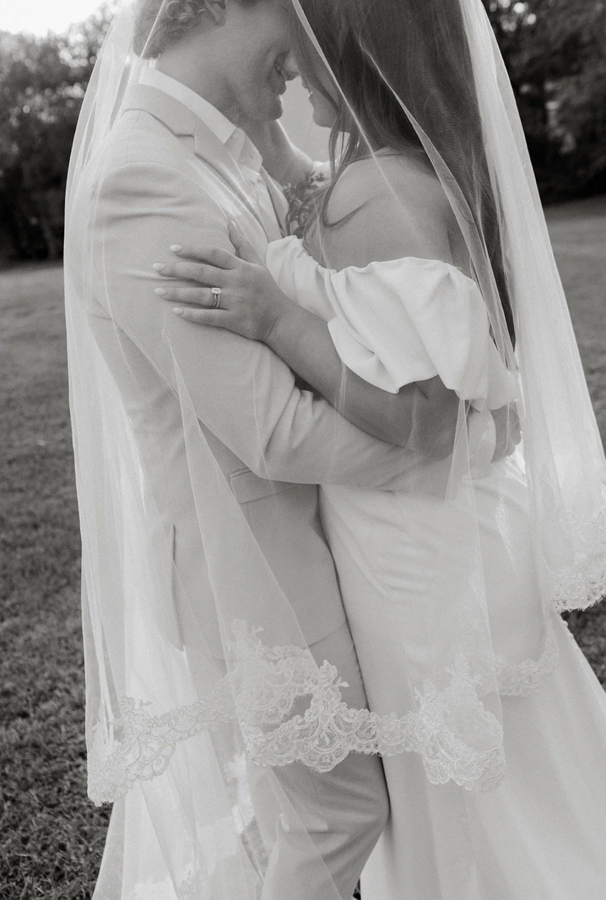 A bride and groom embrace closely under a veil during their wedding outdoors.