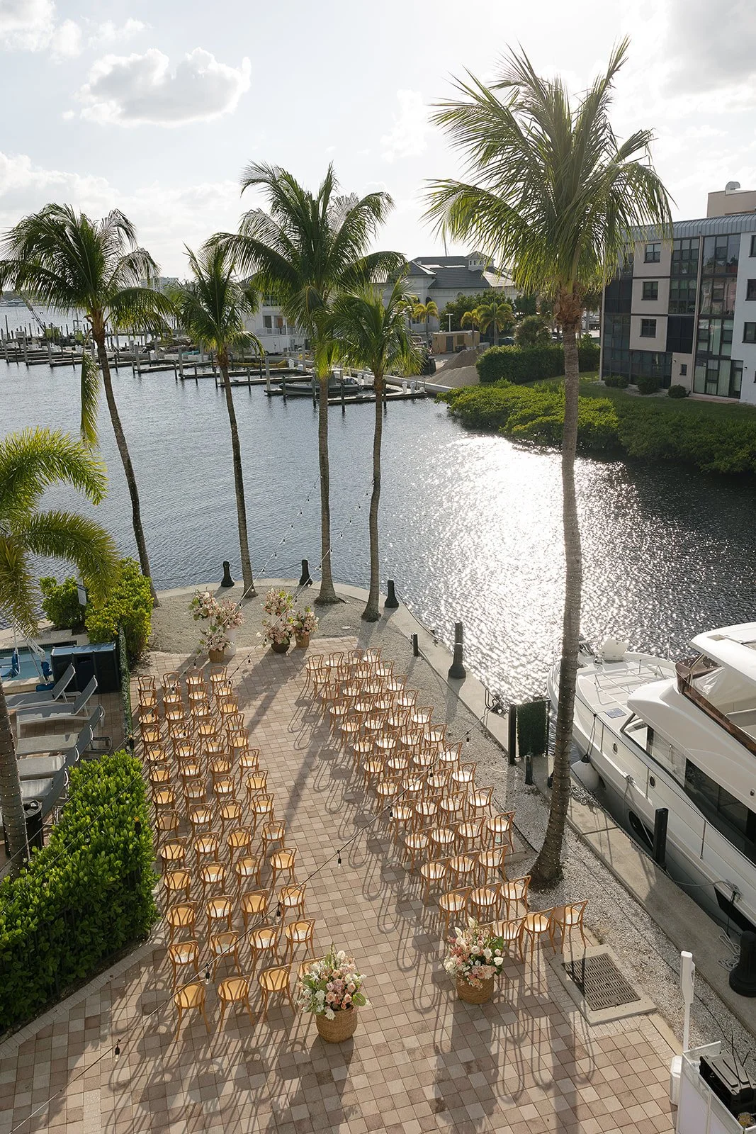 Outdoor event setup along a waterfront with chairs arranged in rows, large flower pots, palm trees, and a docked yacht.