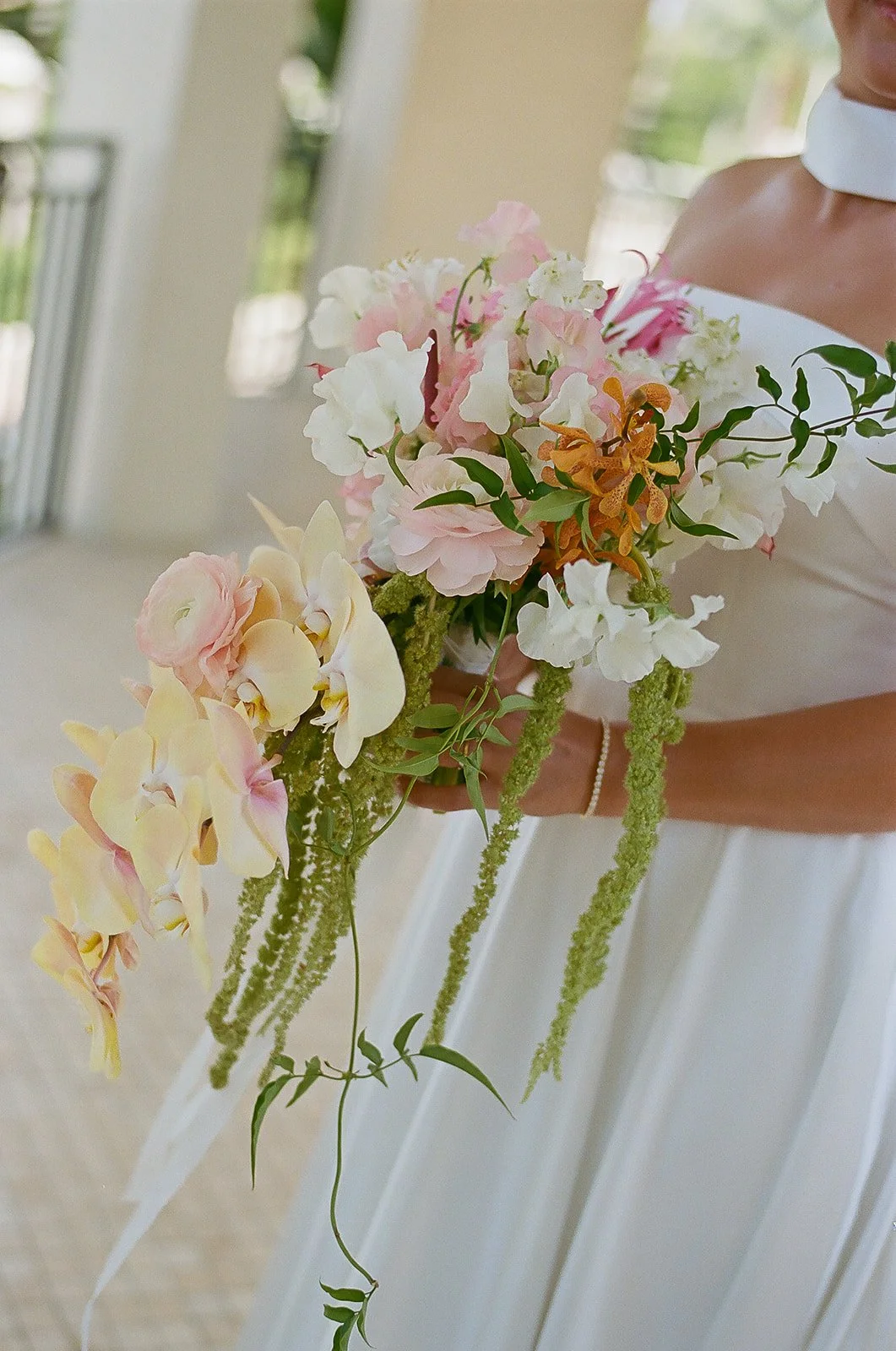 A person in white holding a cascading bouquet of pink, white, yellow, and orange flowers with greenery, standing in a bright outdoor setting.