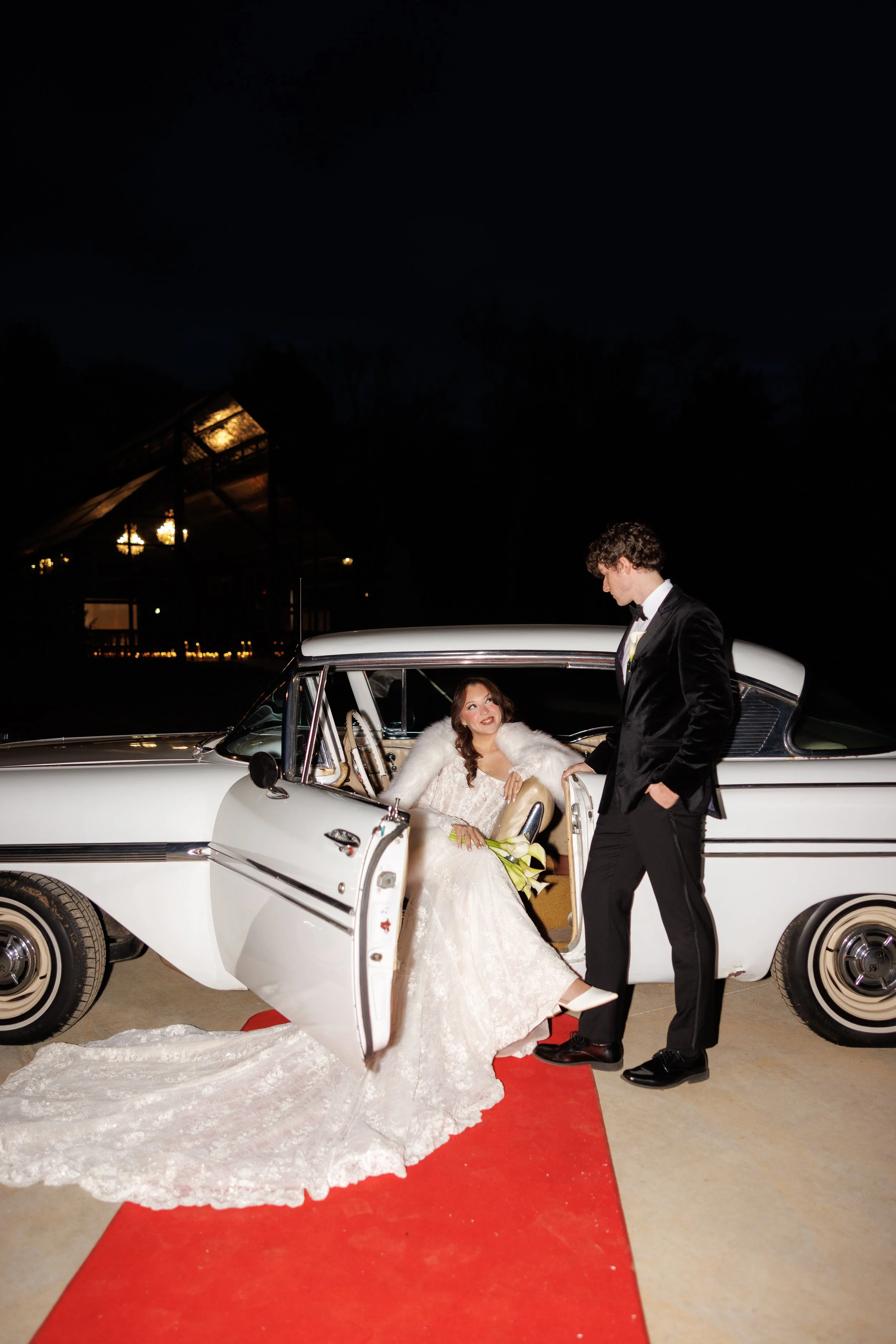 A bride in a white lace wedding dress and fur shawl sitting in a classic white car with the door open, holding a bouquet, as a groom in a black tuxedo stands beside her holding her hand on a red carpet at night.