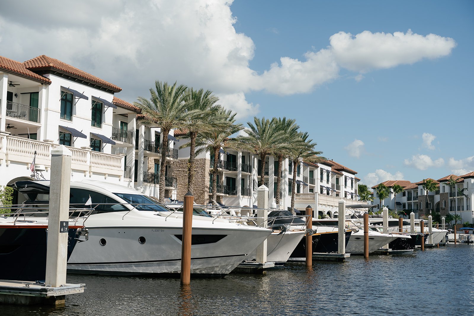 Luxury yachts docked at a marina with modern white buildings and palm trees in the background under a partly cloudy sky.