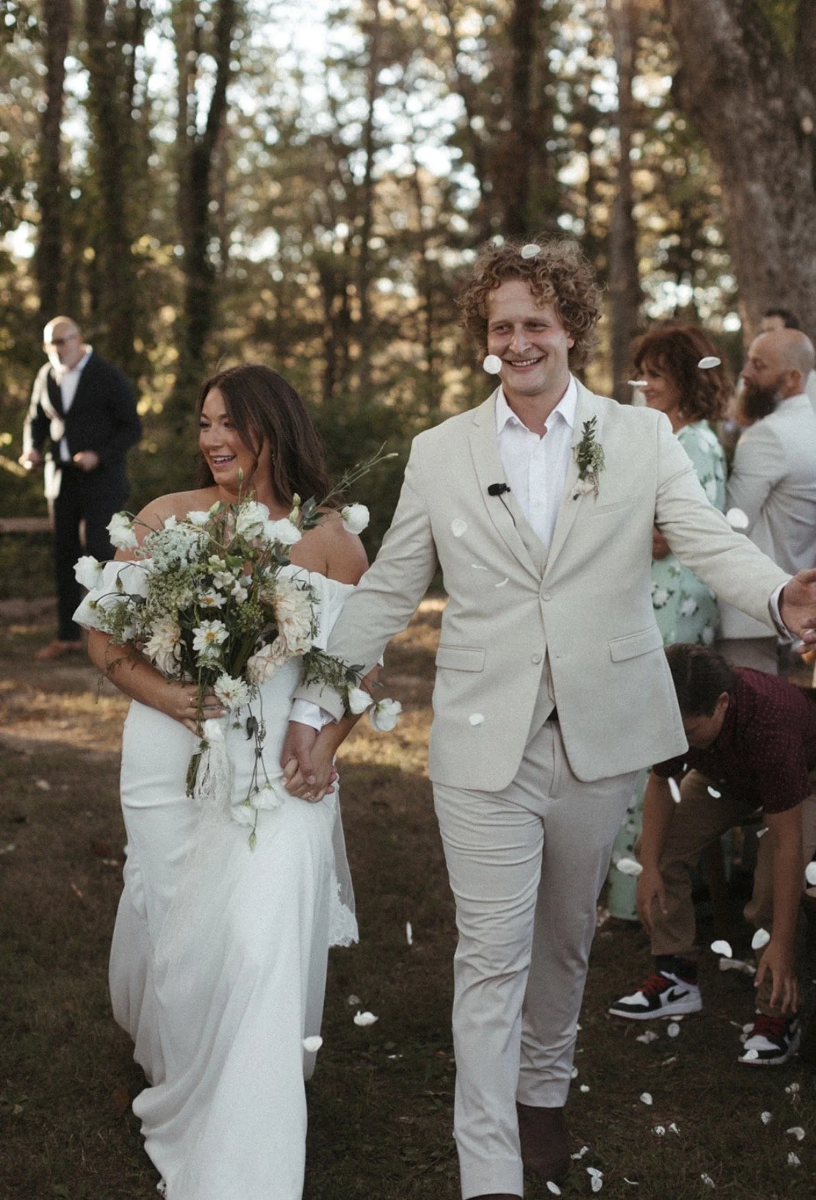 A newlywed couple walking outdoors during their wedding celebration, holding hands, with guests in the background and flower petals falling.