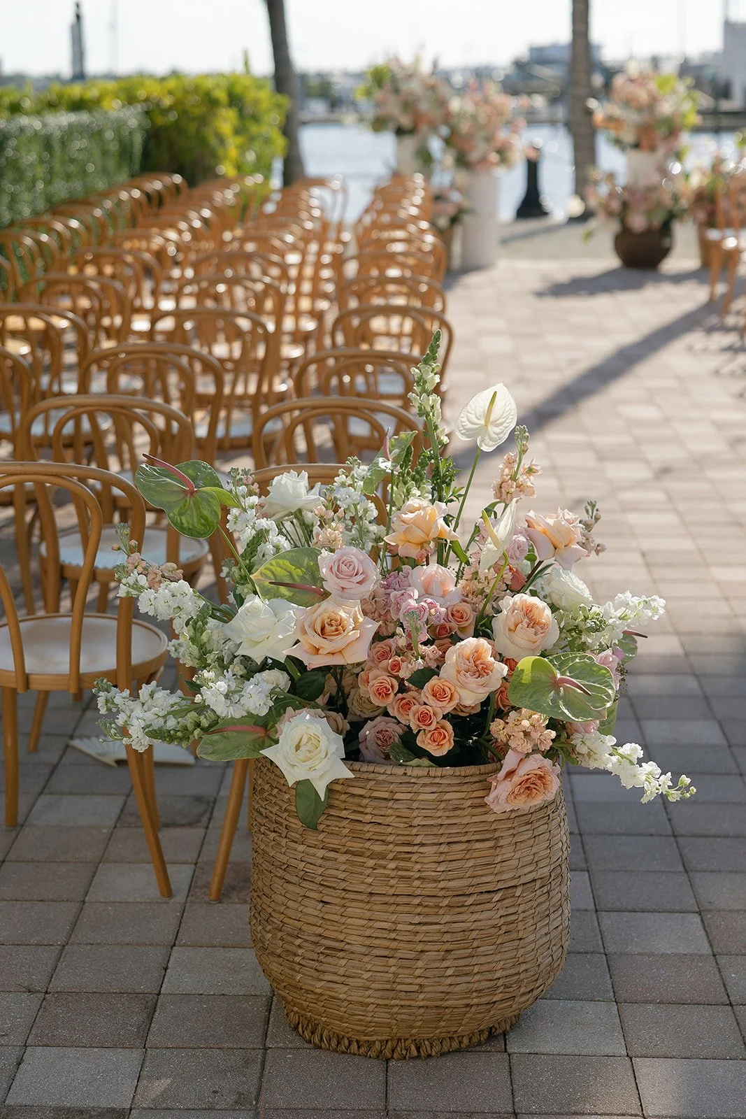 A large woven basket filled with pink, white, and peach roses, white anthuriums, and greenery in an outdoor setting with empty chairs and floral arrangements along a waterfront walkway.