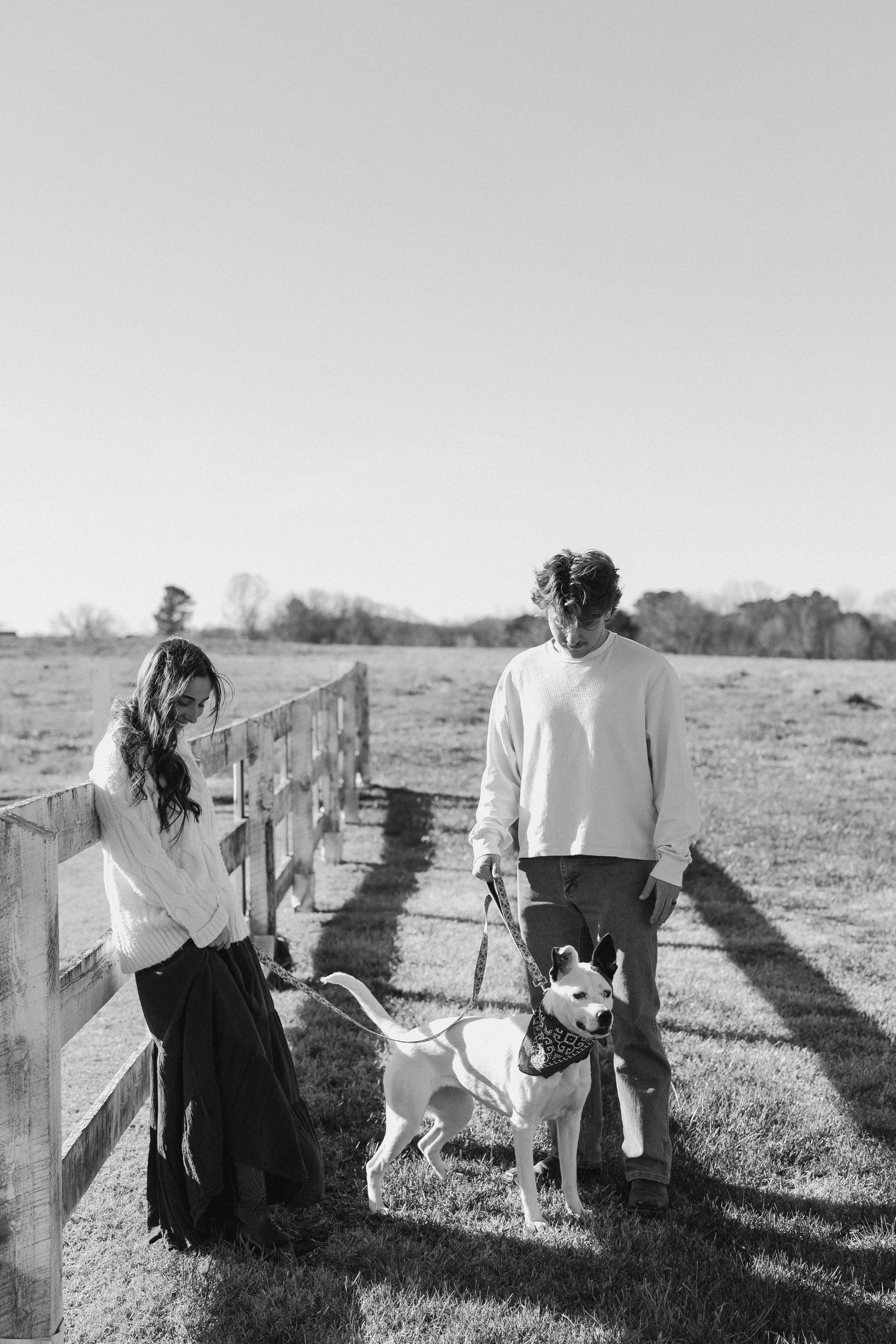 A black and white photo of a young man and woman walking a dog on a grassy field with a wooden fence on the left side. The woman is leaning on the fence, smiling, while the man holds the dog on a leash. The dog is wearing a bandana and looking at the camera.