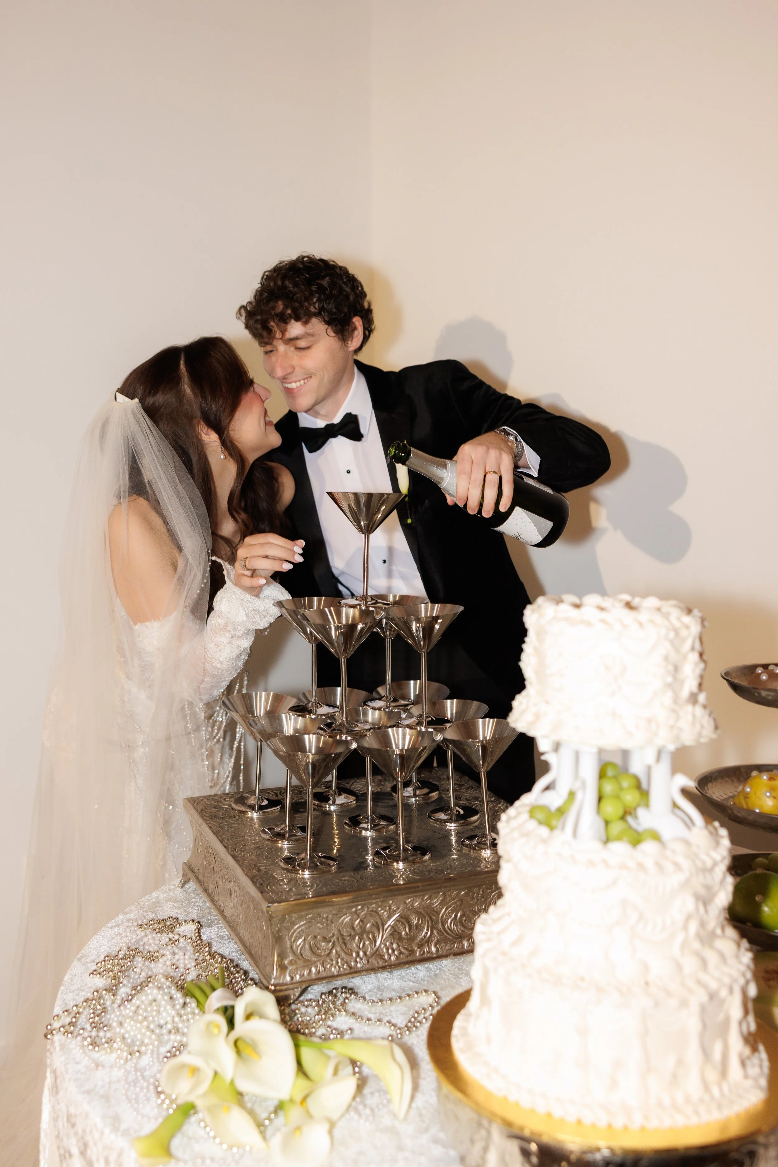 A newlywed couple at their wedding celebration pouring champagne into a pyramid of glasses, with a wedding cake and floral decorations in the foreground.