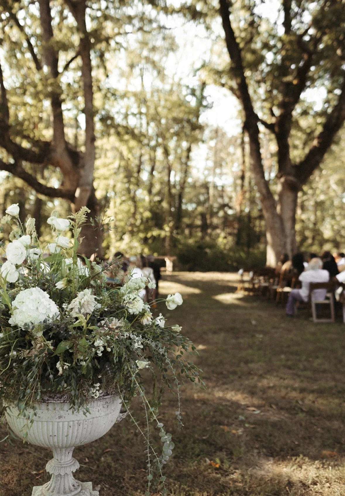 A white floral arrangement in a pedestal vase at an outdoor event, with guests seated in rows of chairs under tall trees with green leaves.