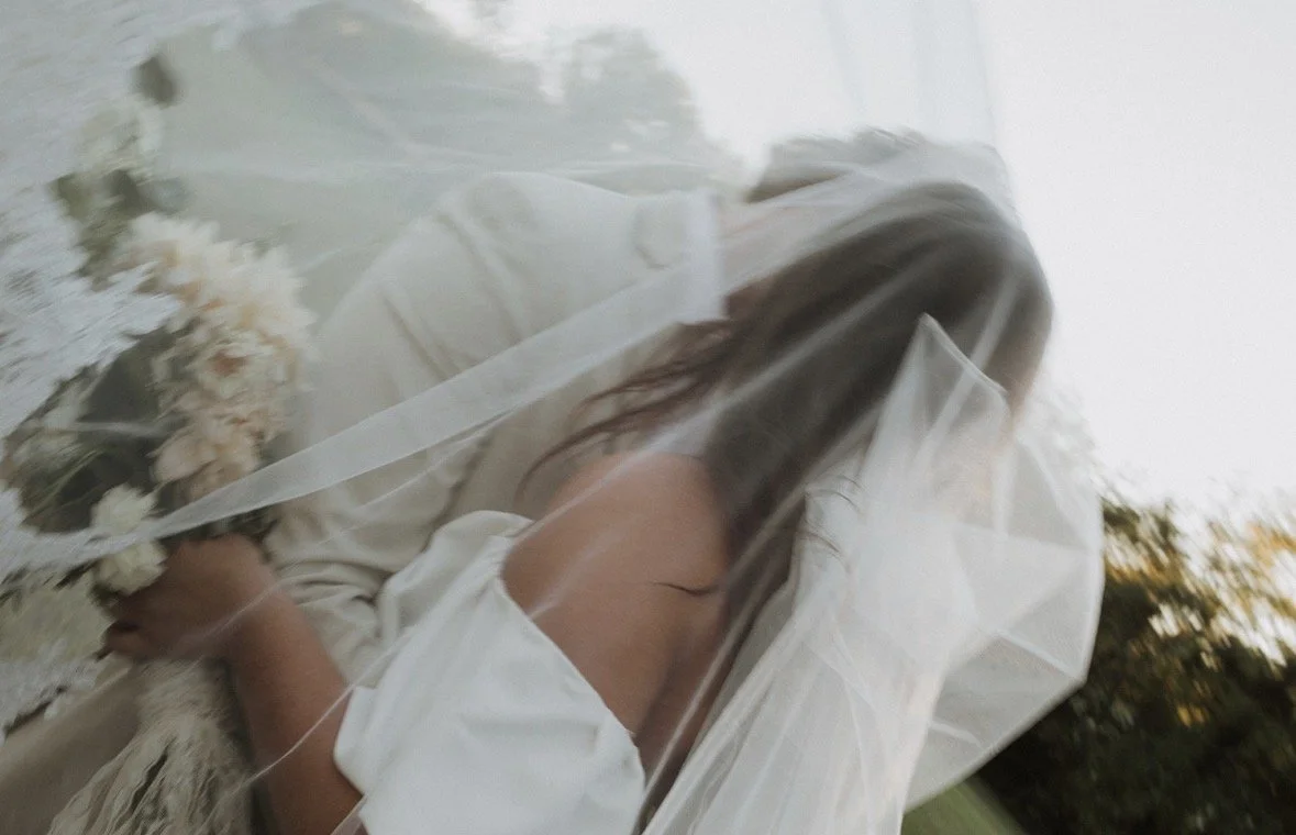 A woman in a white dress with a veil holding a bouquet of flowers, seen through a glass surface, likely at a wedding or bridal event, outdoors with trees in the background.