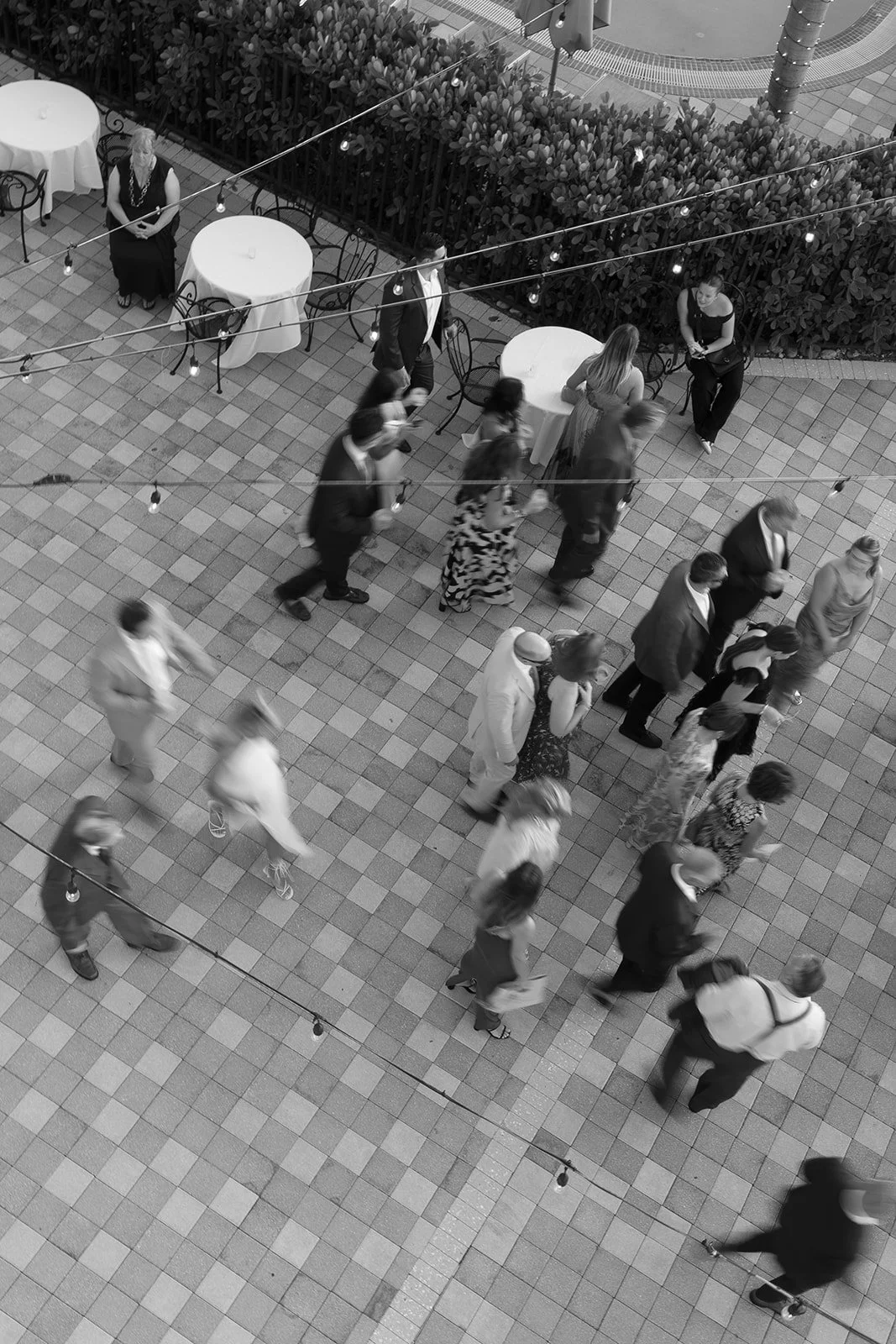 Black and white photo of a group of people standing in line at an outdoor event, with some tables and string lights overhead.