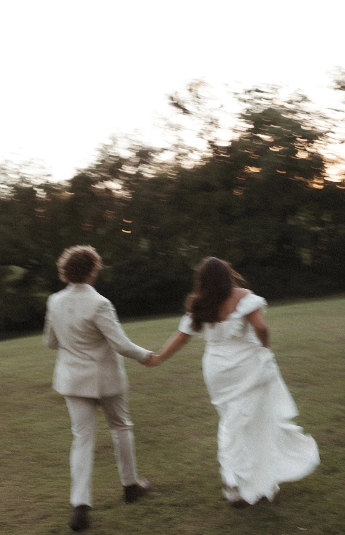 A couple holding hands and running across a grassy field, with trees in the background and a sunset sky.