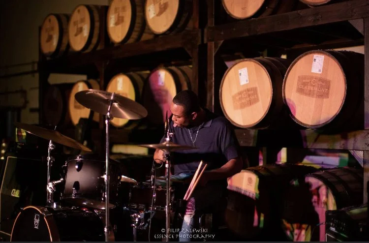 A person playing drums in a room with wooden barrels stacked on shelves in the background.