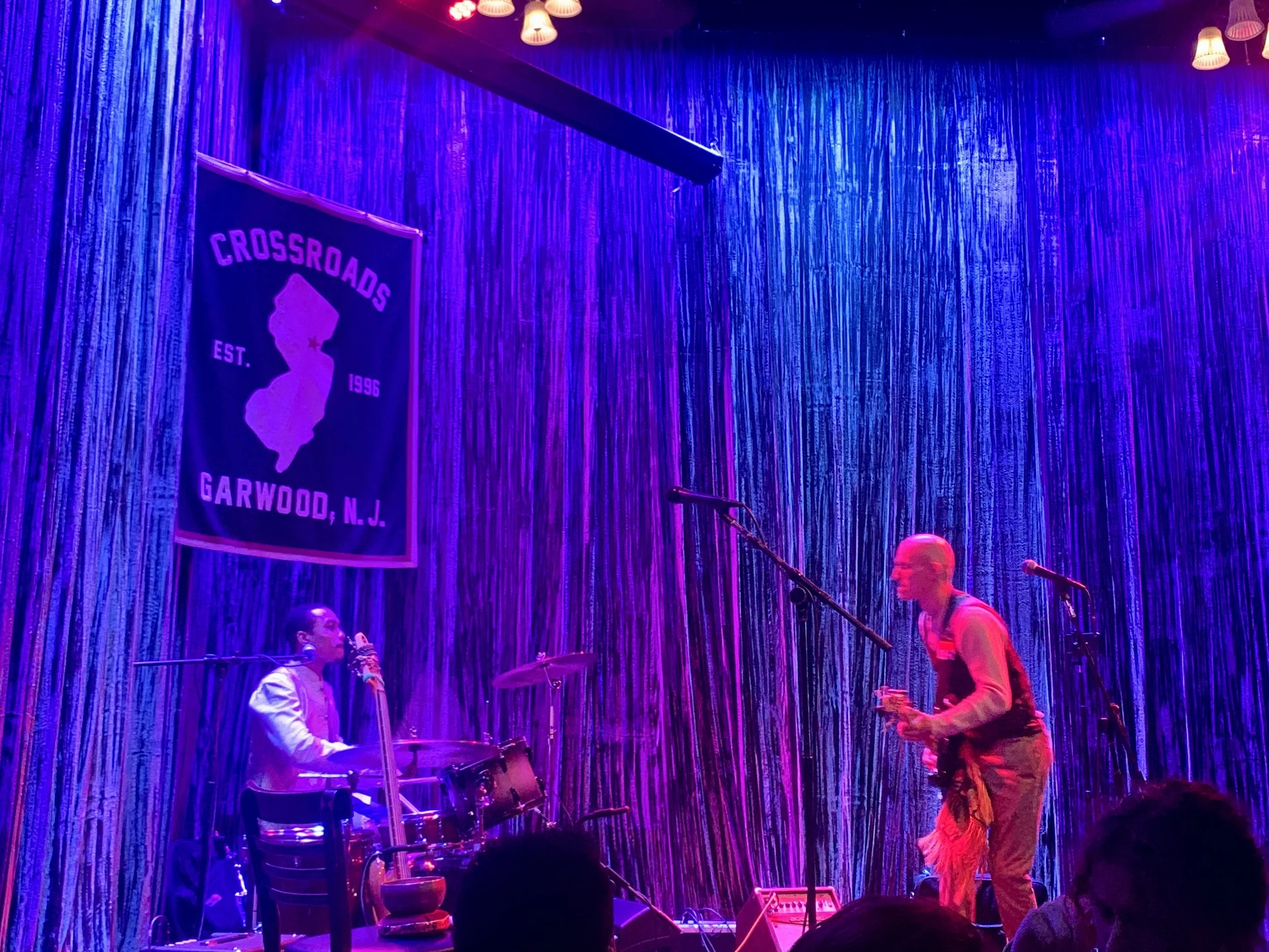 Two musicians performing on stage with vibrant purple and blue lighting, one playing drums and the other playing a wind instrument. A banner on the wall reads 'Crossroads, Est. 1996, Garwood, N.J.'