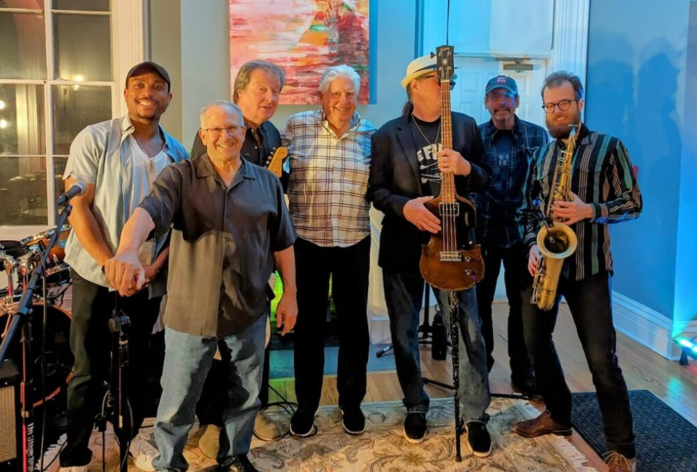 Group of eight musicians posing in a room, some holding instruments including a guitar, saxophone, and bass, with a colorful painting on the wall behind them.