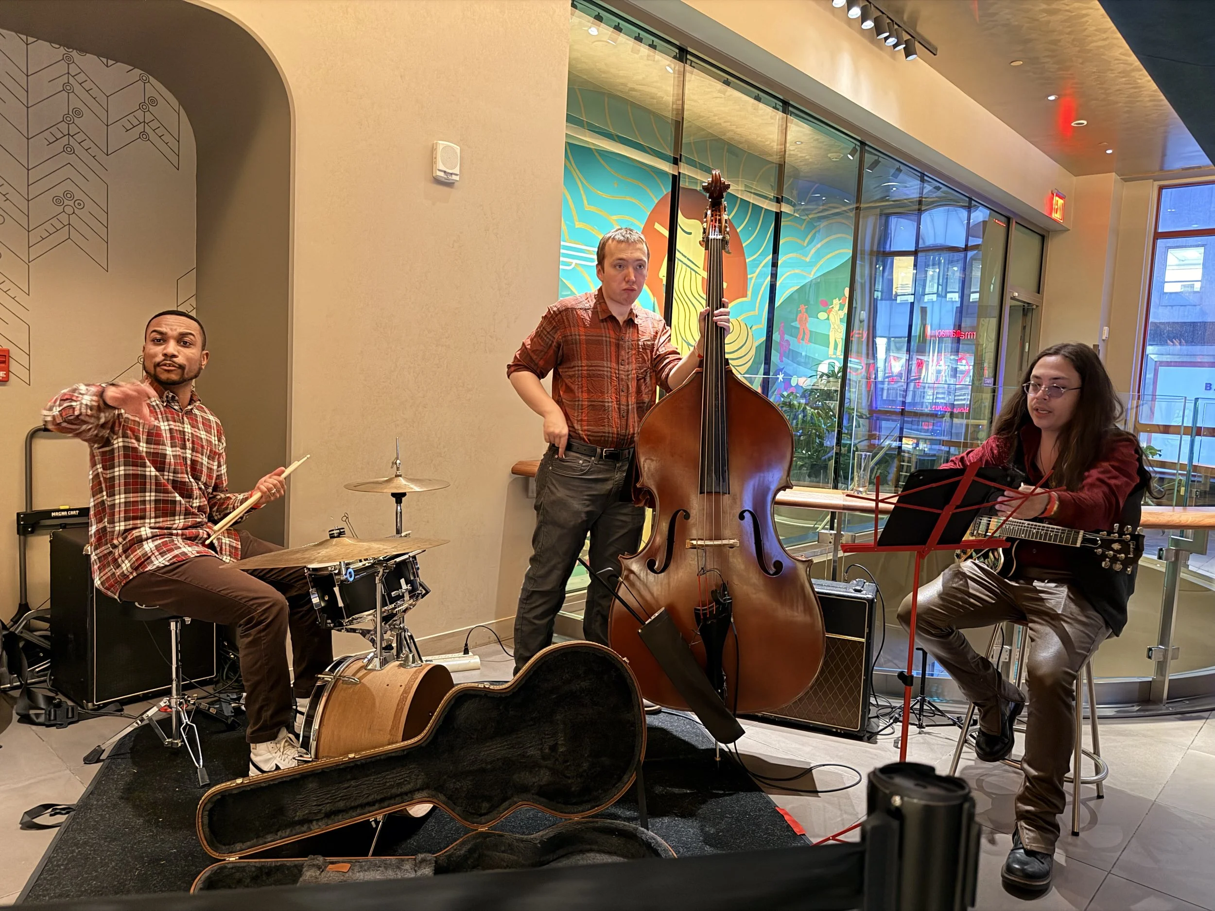 A jazz band performing indoors with three musicians: a drummer on the left, a double bassist in the center, and a guitarist on the right. They are playing their instruments on a small stage with a colorful mural behind them and large glass windows sh