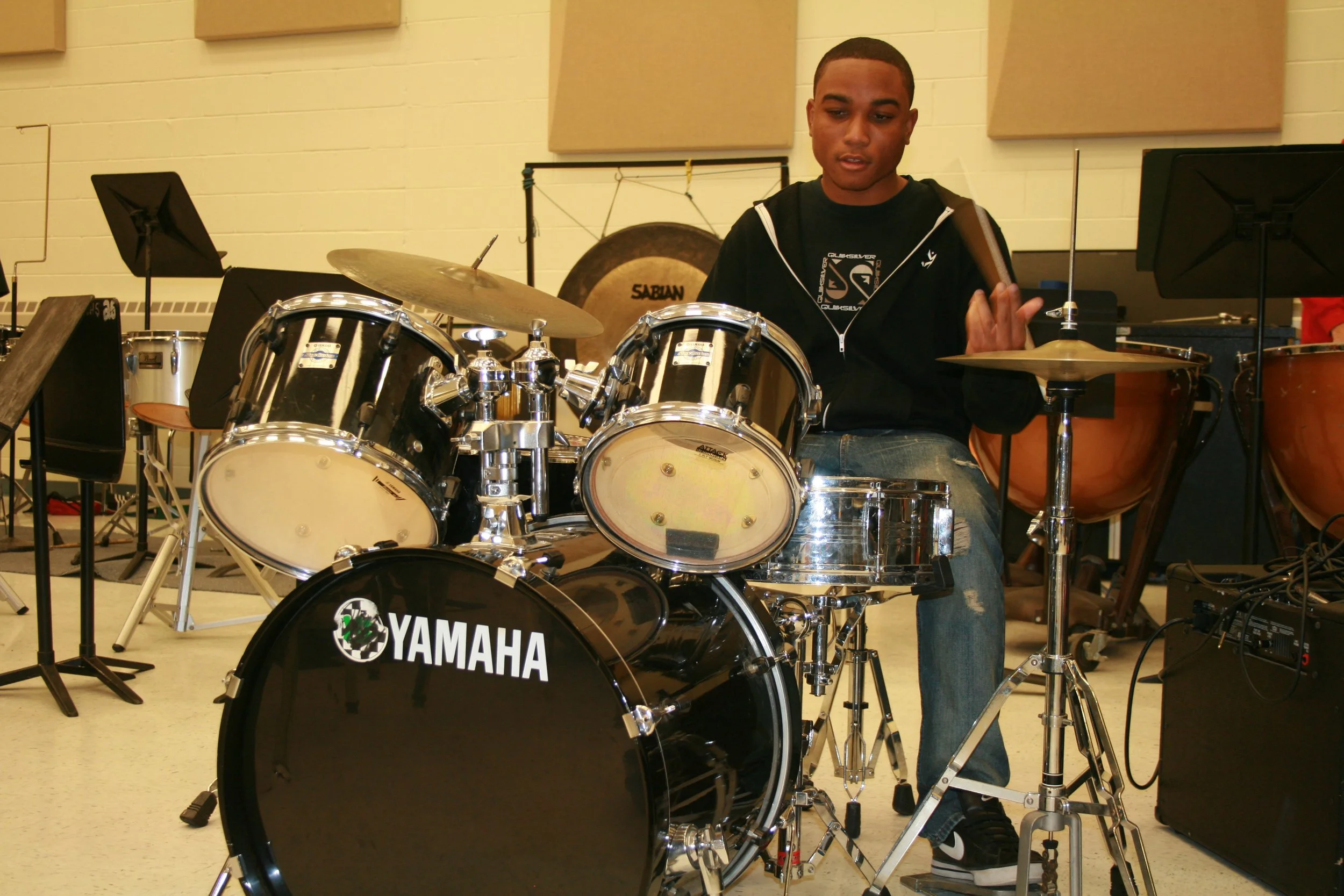 A young man playing a Yamaha drum set in a music room with other percussion instruments and music stands.