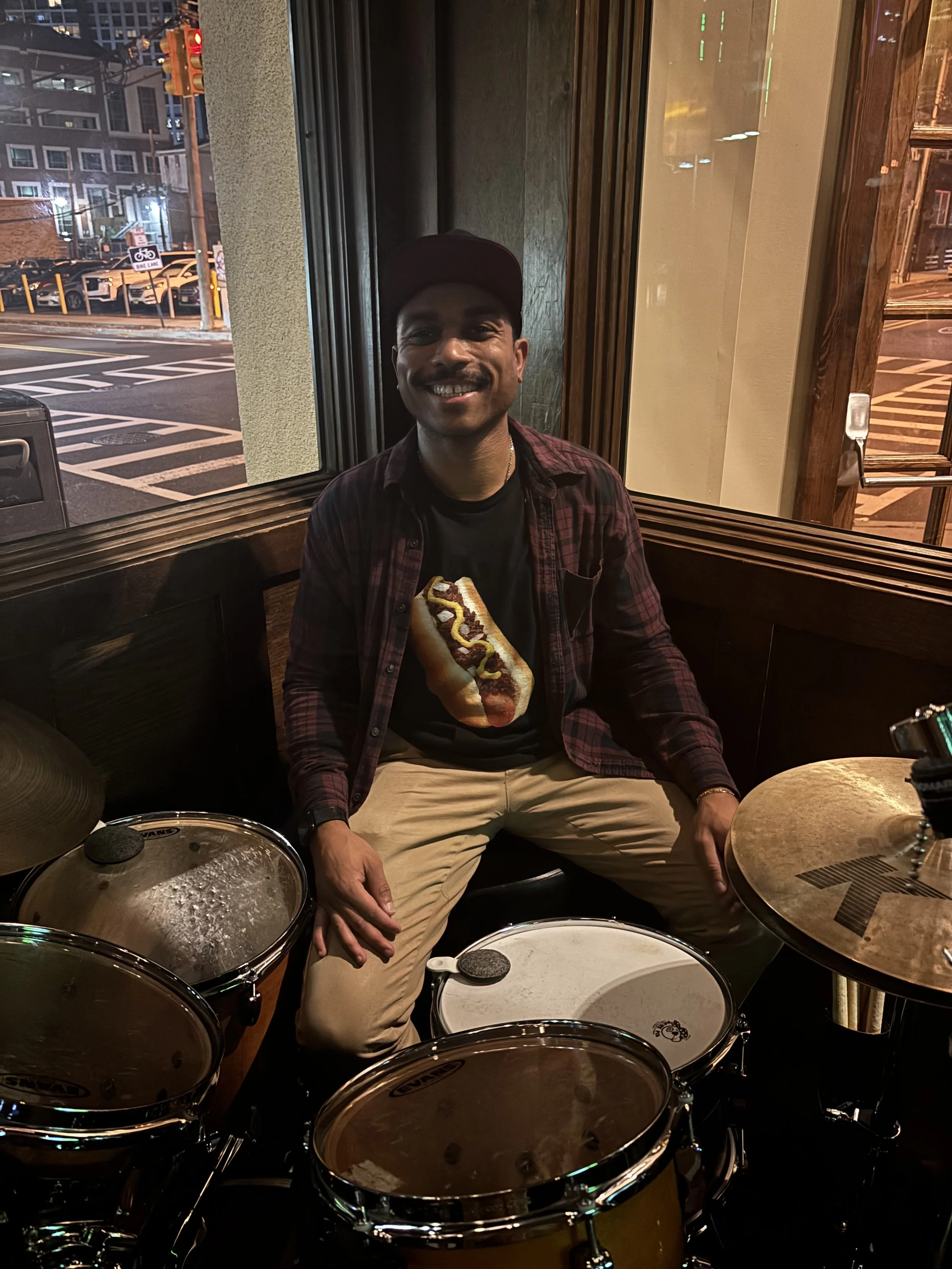 A man sitting inside a restaurant, smiling at the camera, wearing a black t-shirt with a hot dog graphic, a plaid shirt, and a black cap, surrounded by a drum set.