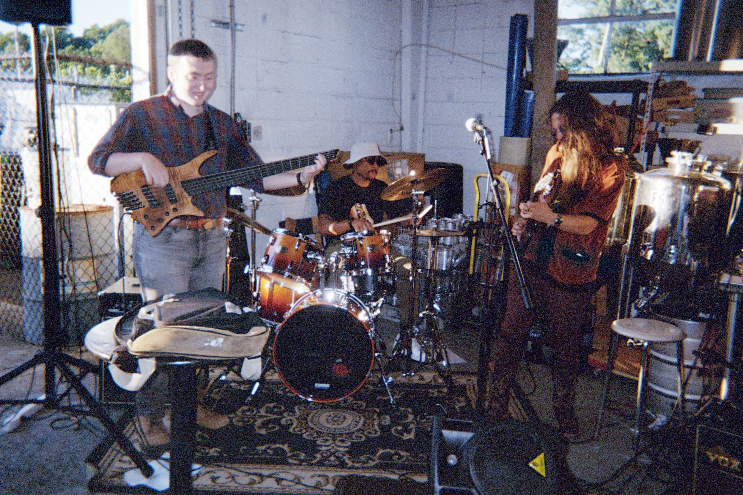 A three-member band rehearsing in a garage, playing various instruments including a bass guitar, a drum set, and a guitar. The garage has a window with a view of trees outside, and the scene is lit with natural light.