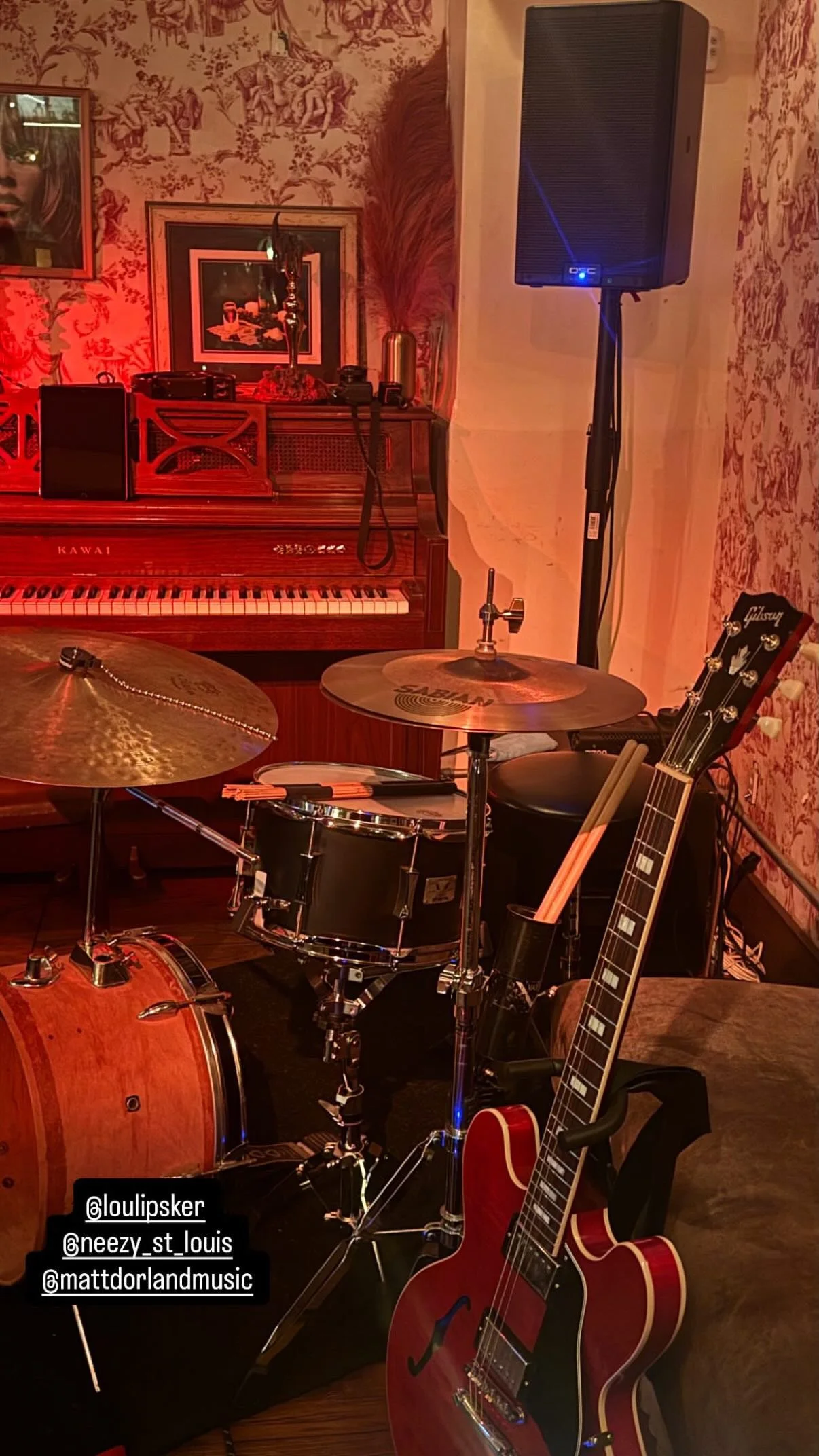 A music room with a red electric guitar, a drum set, a keyboard, and a speaker, with ornate wallpaper and framed pictures on the wall.
