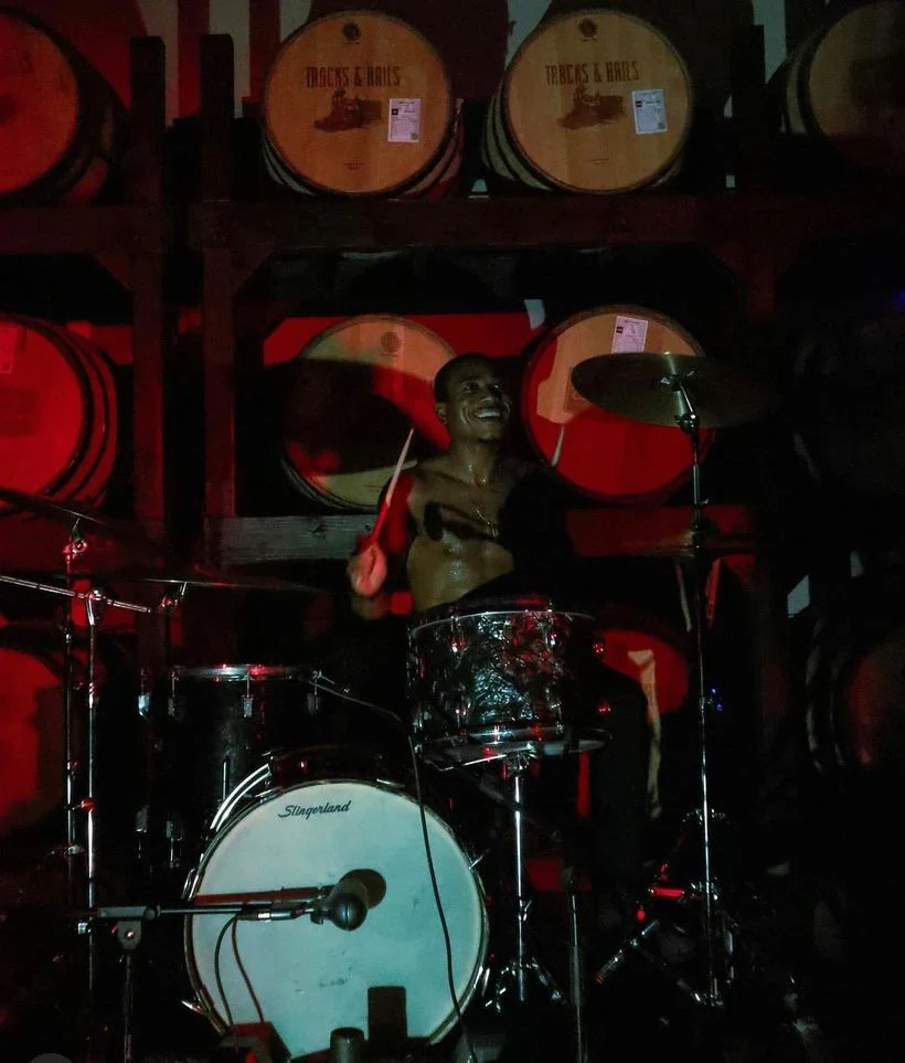 A shirtless man playing drums on a stage with barrels in the background.