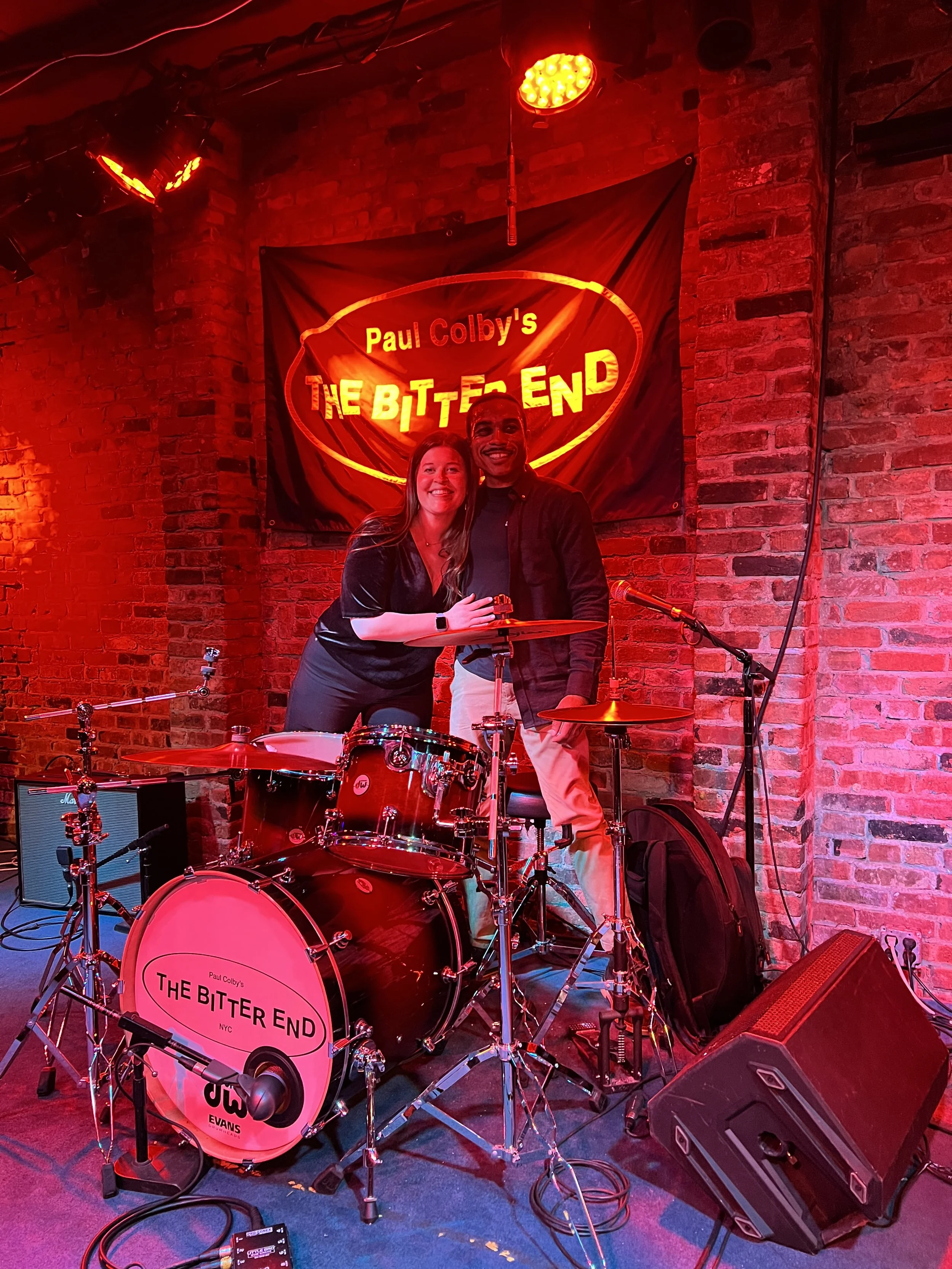 Two people smiling on stage behind a drum set with a brick wall and a banner reading "Paul Colby's The Bitter End" in the background.