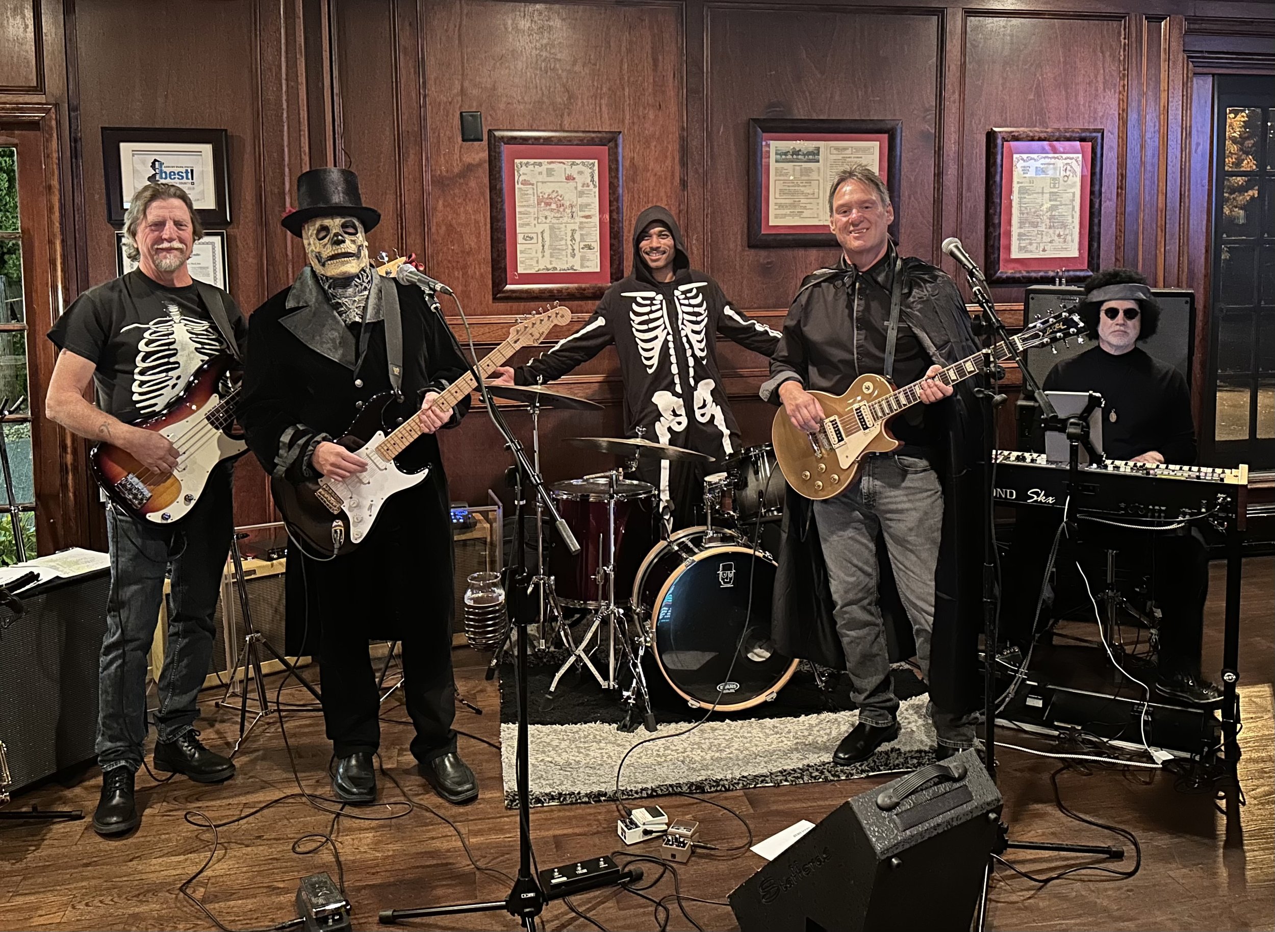 Five musicians in skeleton-themed costumes performing with instruments in a wood-paneled room.