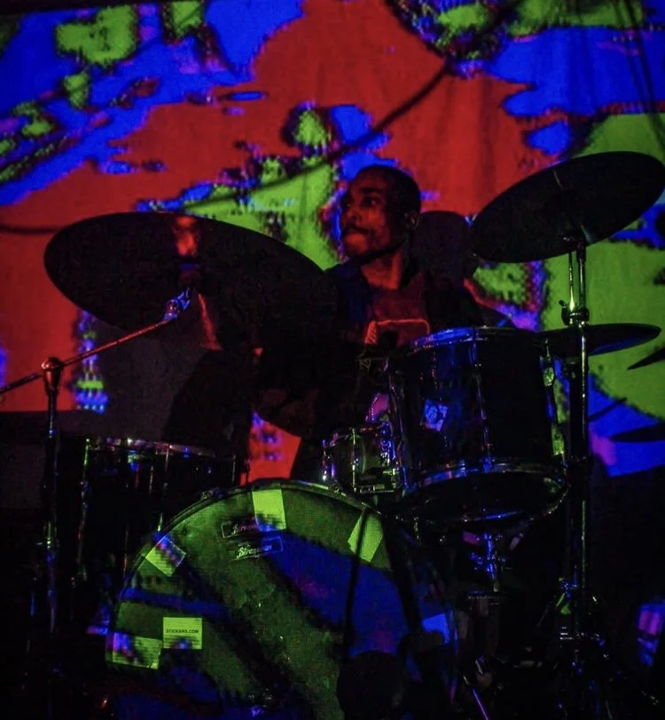 A man playing drums during a live performance with colorful digital visuals projected behind him.