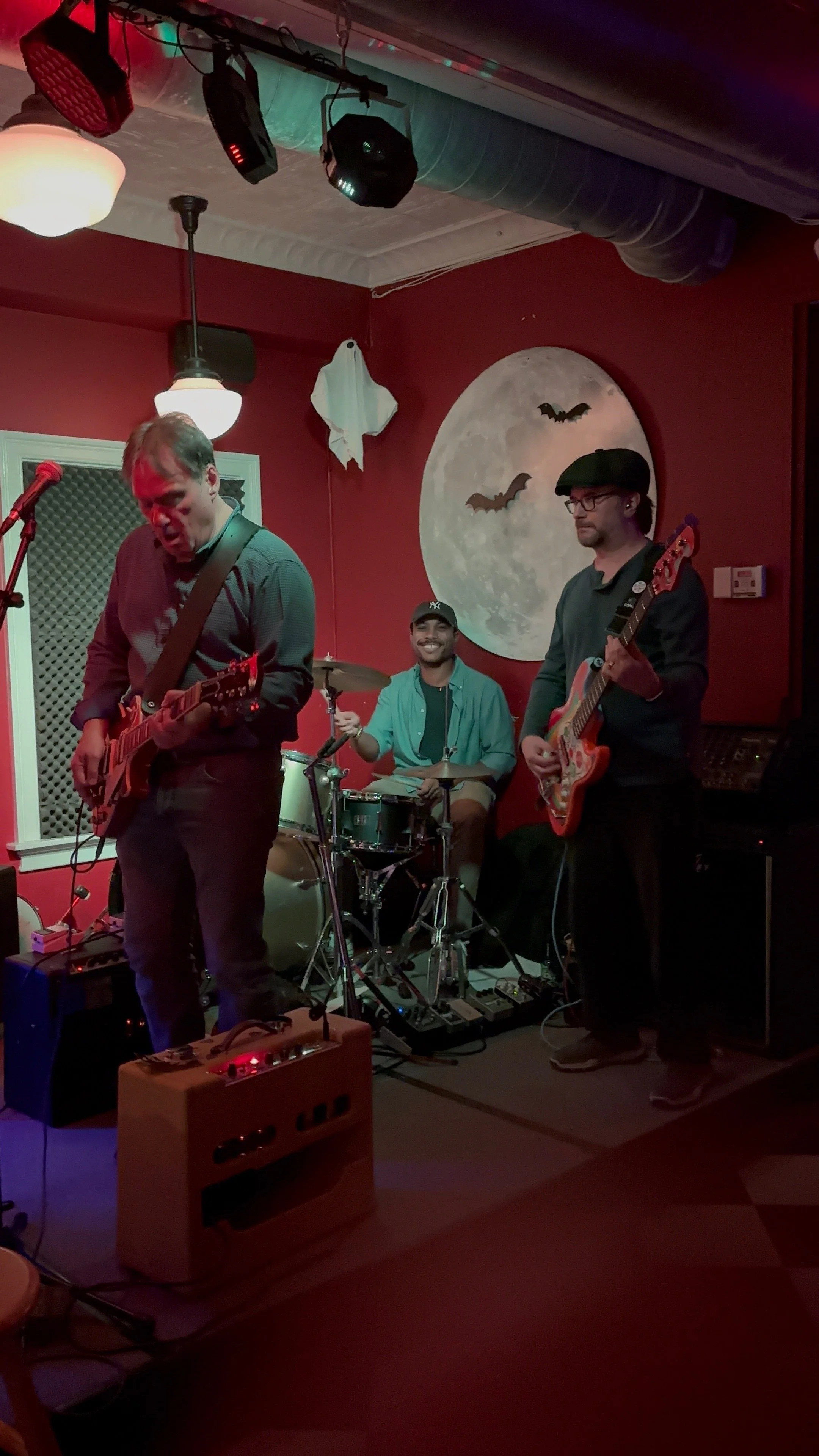 A band performing live with two guitarists and a drummer on stage, with a large moon and bats painted on the red wall behind them.