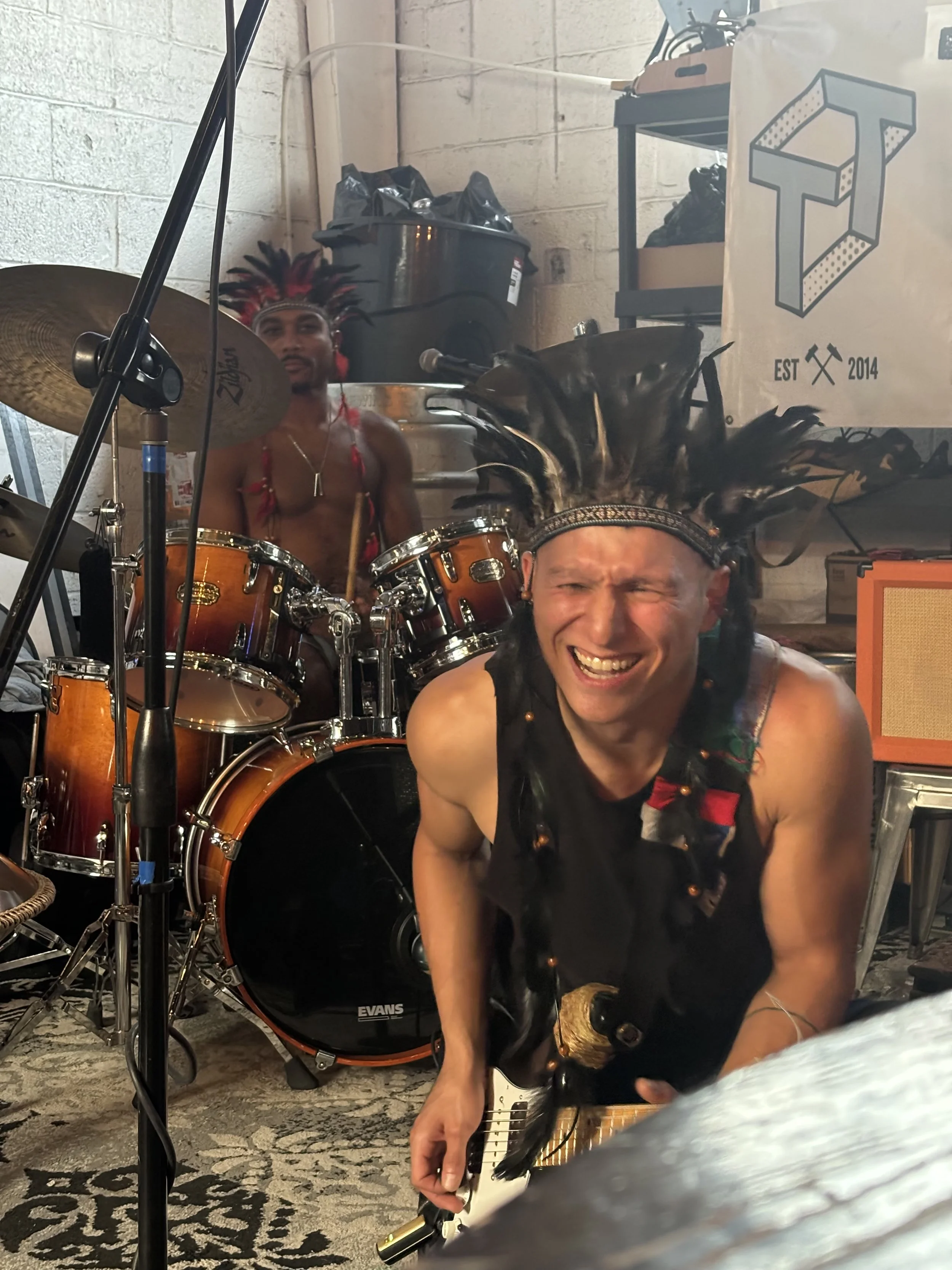 Two men are in a room with musical instruments. The man in the foreground is smiling, wearing a feathered headdress, and holding a guitar. The man in the background is sitting behind a drum set, wearing a feathered headpiece, and looking at the camer