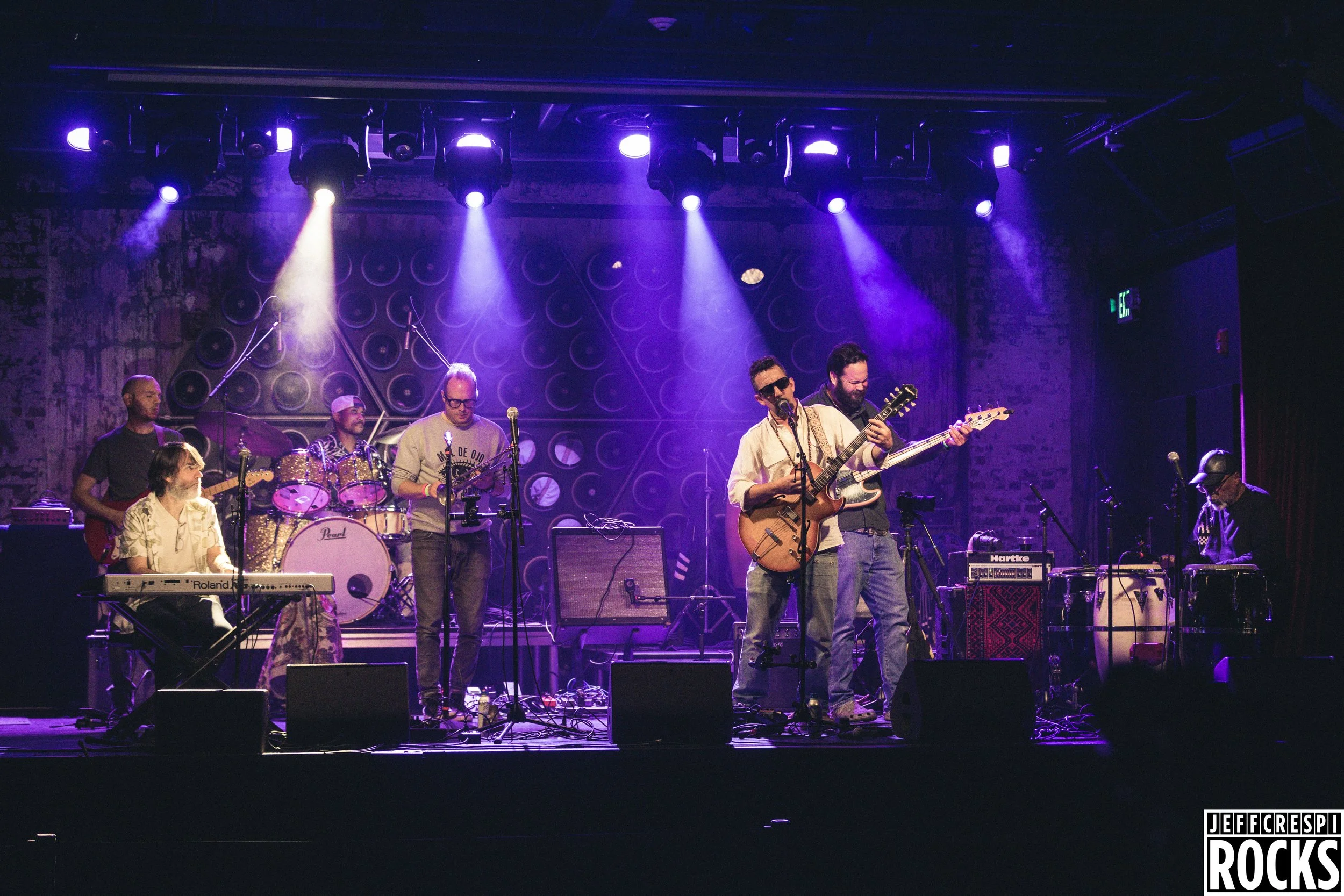 A band performs on stage with purple and white lighting, featuring musicians playing keyboards, drums, electric guitar, acoustic guitar, and other instruments, in a venue with brick walls and circular sound equipment on the backdrop.