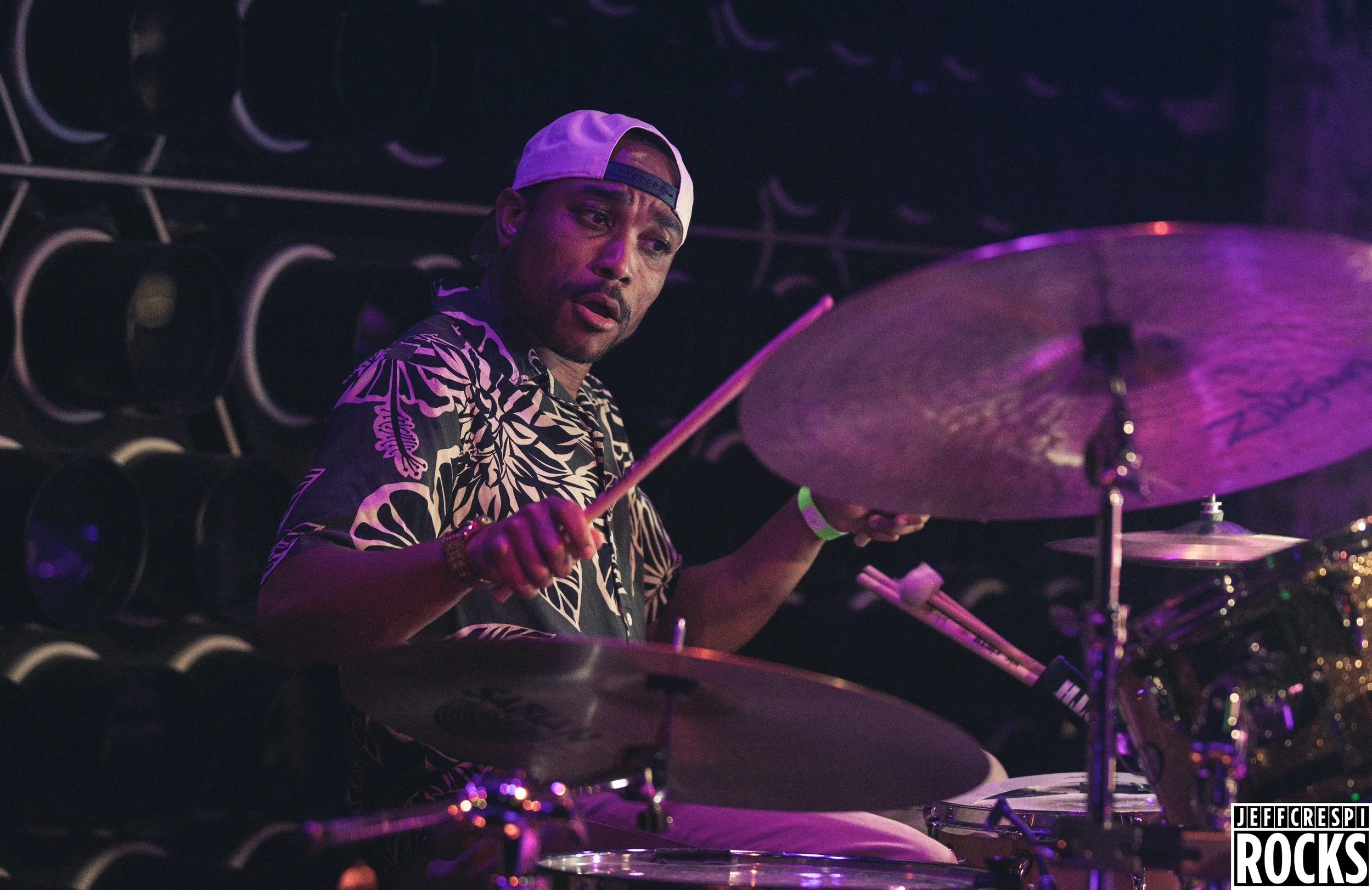 A man playing drums on stage under purple lighting, wearing a white baseball cap backward and a patterned shirt, with a focused expression.