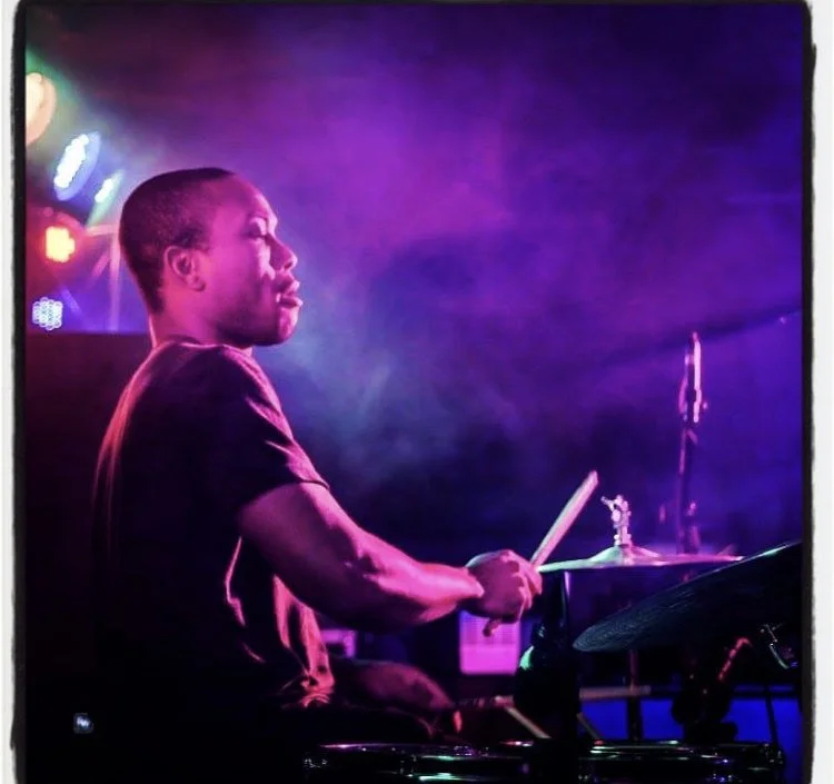 A person playing drums on stage, illuminated by purple and pink stage lights with smoky atmosphere.