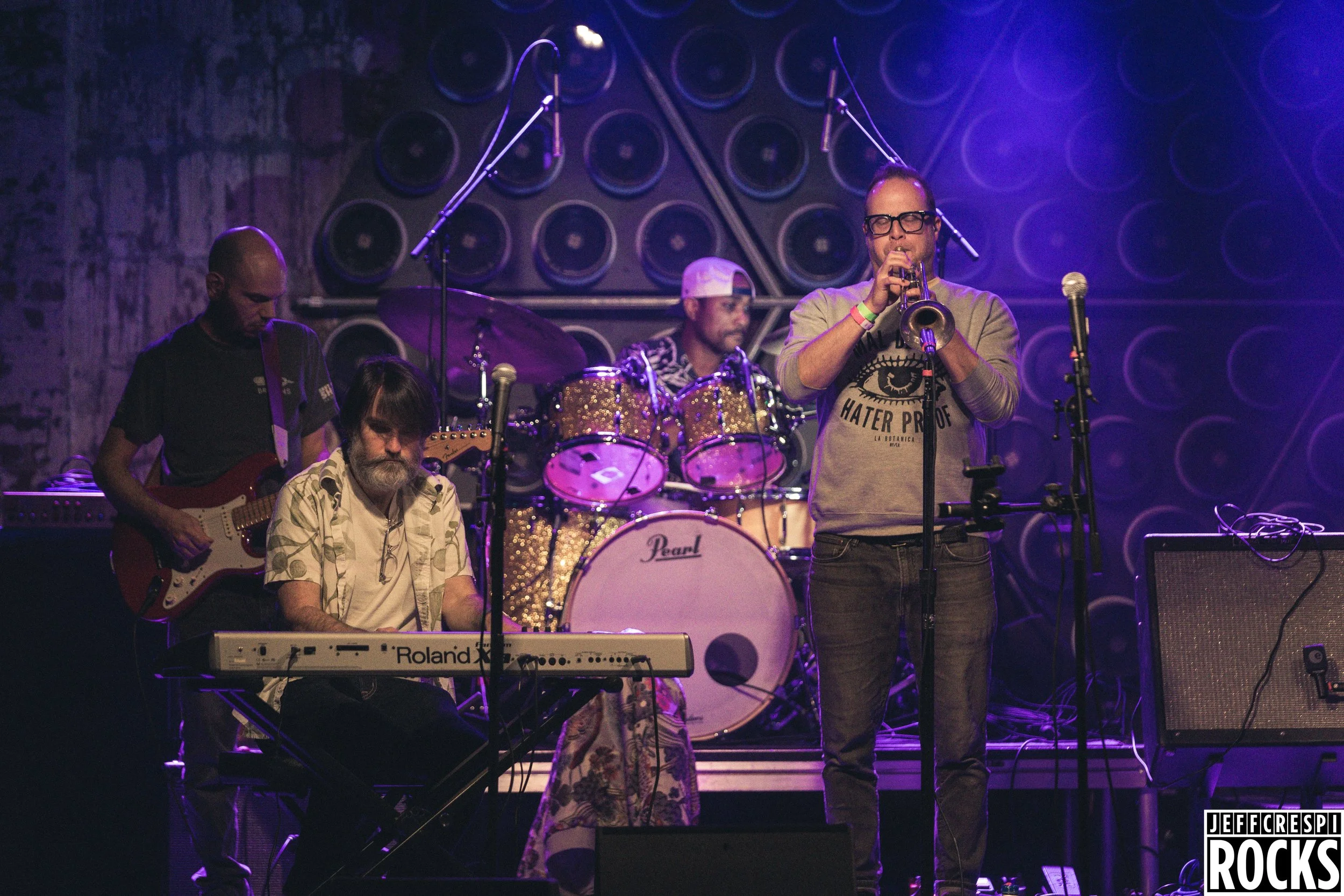 A live music performance featuring four band members on stage. One person plays the keyboard, another on the guitar, a third on drums, and a fourth on trumpet. The stage has multiple large speakers mounted on the back wall, and purple stage lighting 