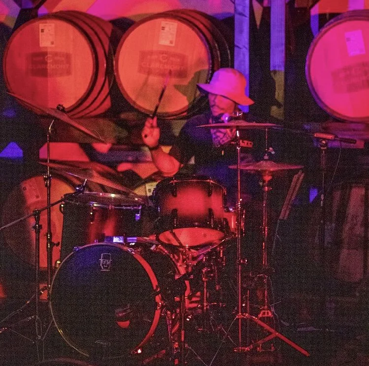 A person wearing a wide-brimmed hat and a face mask playing a drum set in a dimly lit room with red and purple lighting, with large wine barrels in the background.