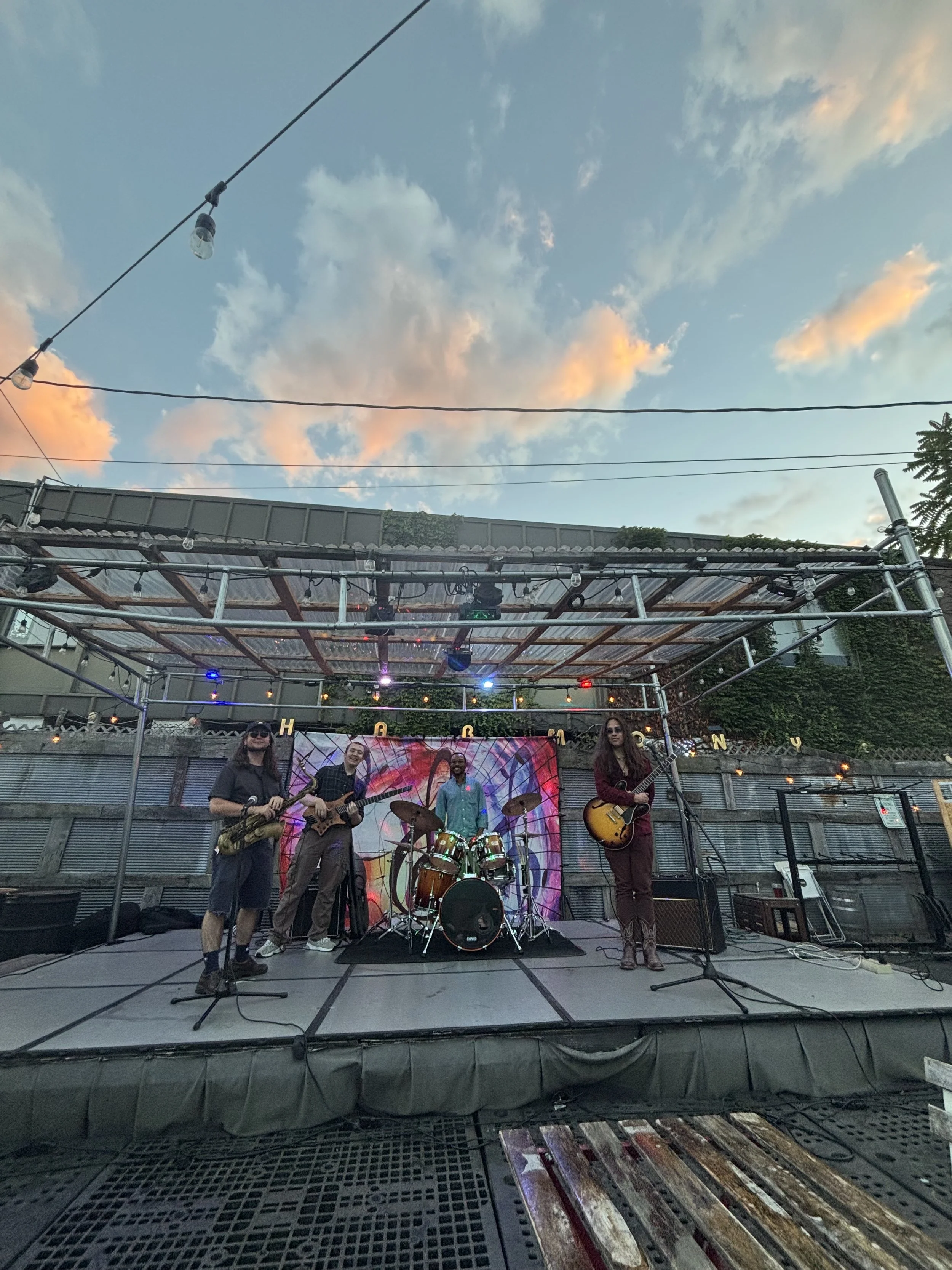 A live outdoor musical performance by a band on a stage with sky and clouds in the background.