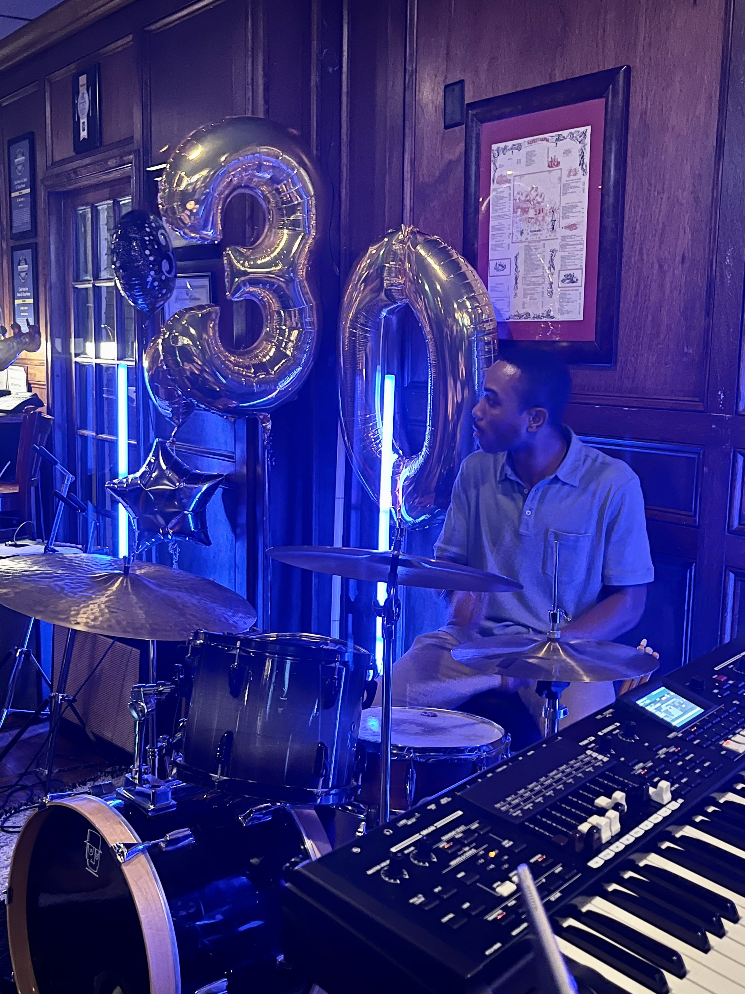 A man sitting behind a drum set and an electronic keyboard at a celebration with silver balloons spelling out '30' and star-shaped balloons, blue lighting, and wooden walls.
