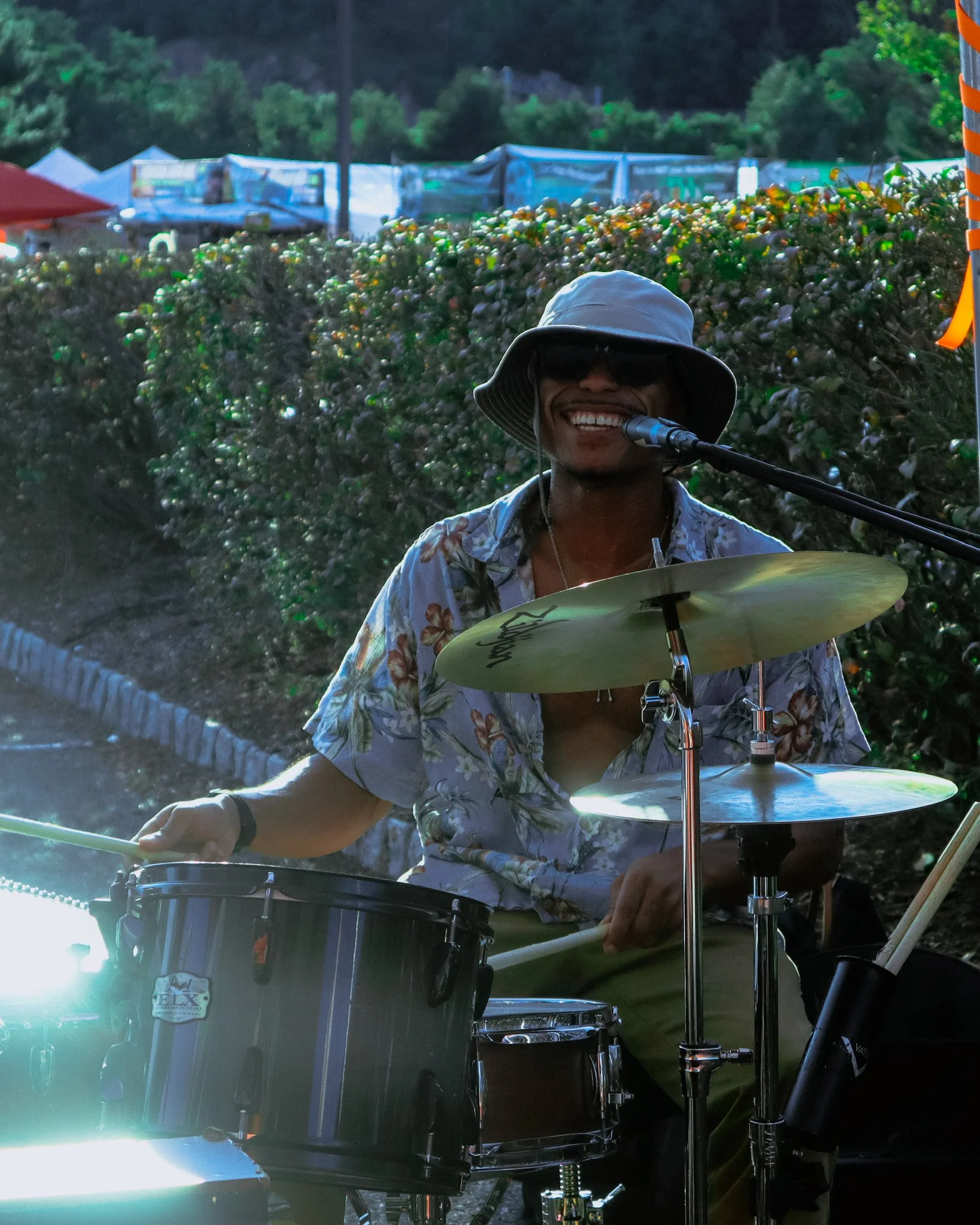 A smiling man wearing sunglasses and a wide-brimmed hat playing a drum set outdoors.
