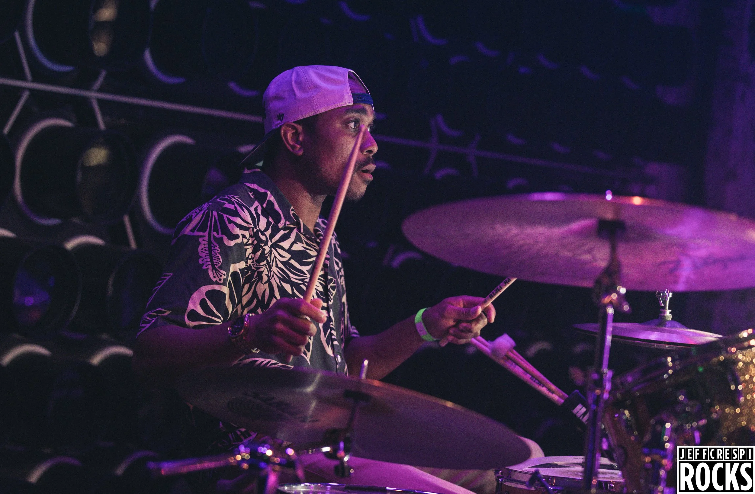 A male drummer wearing a backward cap and floral patterned shirt playing drums on stage under purple lighting.